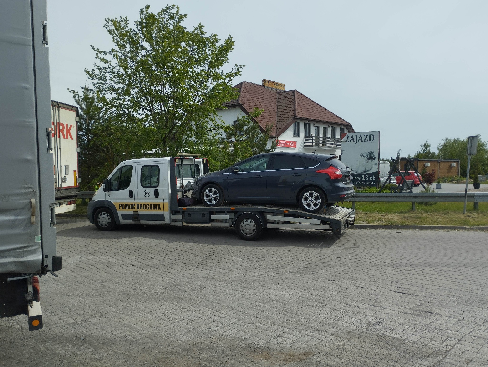 Laweta z szarym Fordem Focus na platformie, na parkingu z kostki brukowej. W tle budynek z brązowym dachem i szyldem Zajazd.