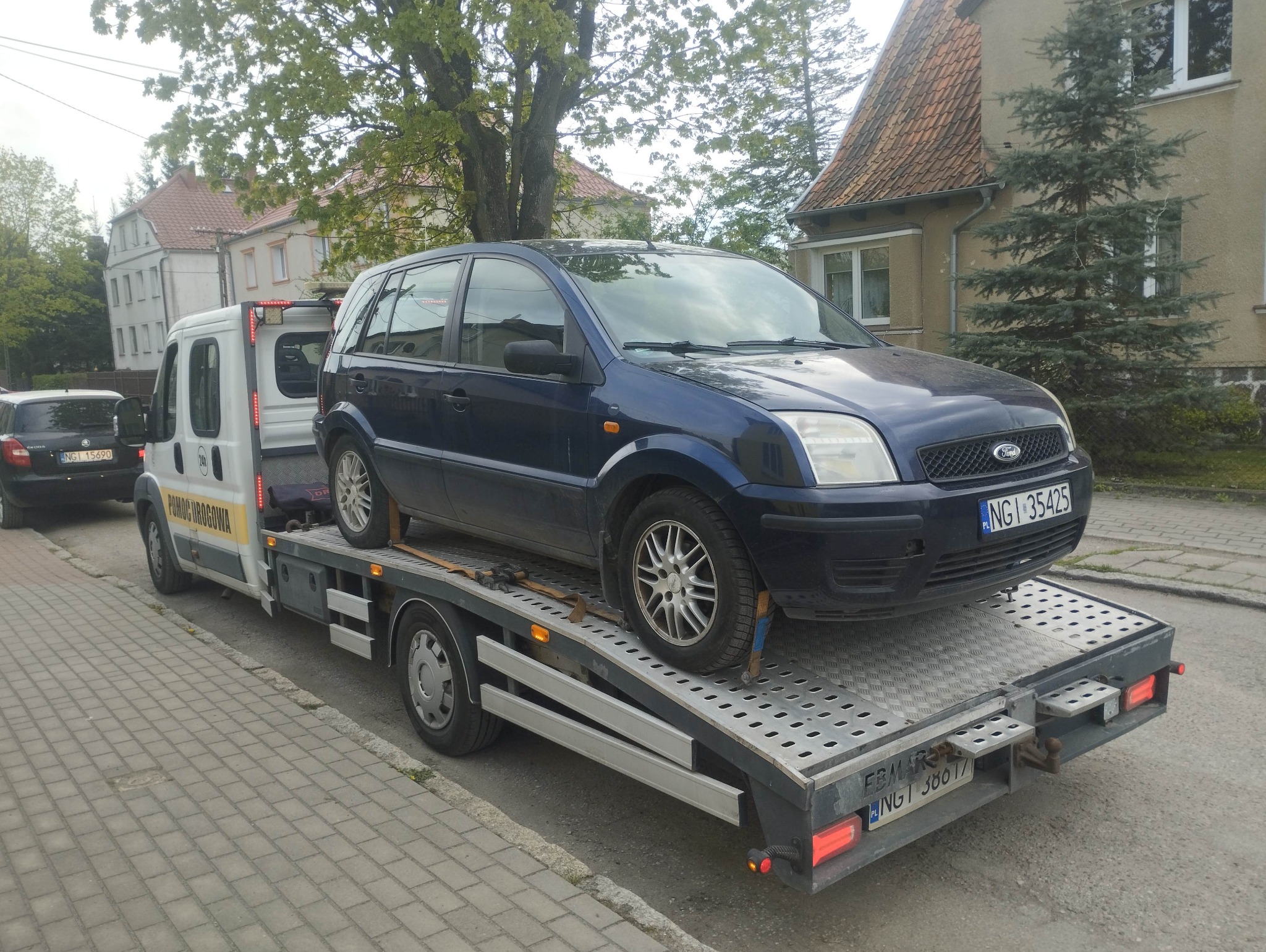 Laweta z niebieskim Fordem Fusion na platformie, tablica rejestracyjna NGI 35425, w tle budynki mieszkalne i drzewa. Pomoc drogowa na osiedlowej ulicy.