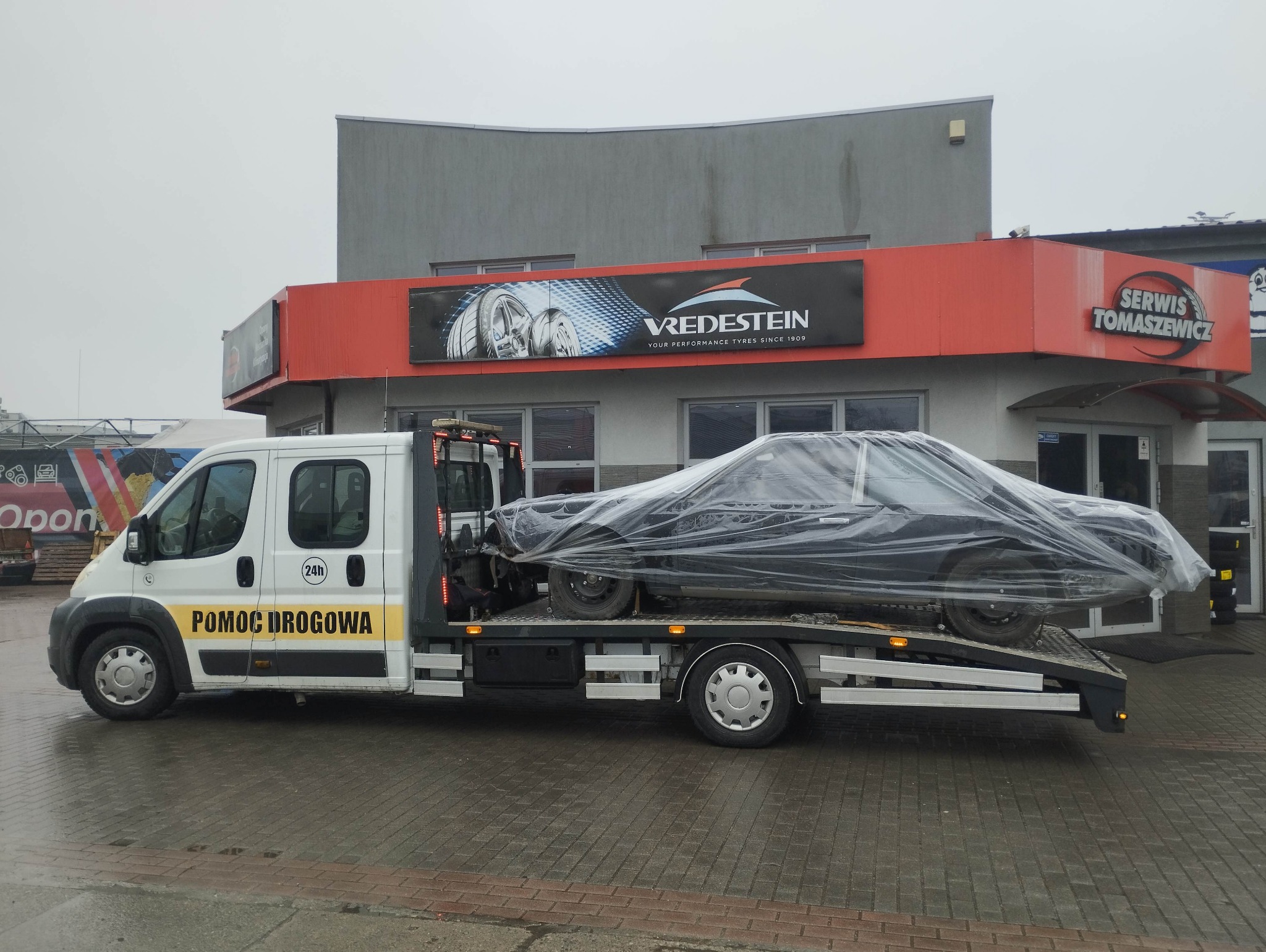 Laweta z uszkodzonym autem pod folią na tle budynku serwisu opon i samochodów. Pomoc drogowa 24h.