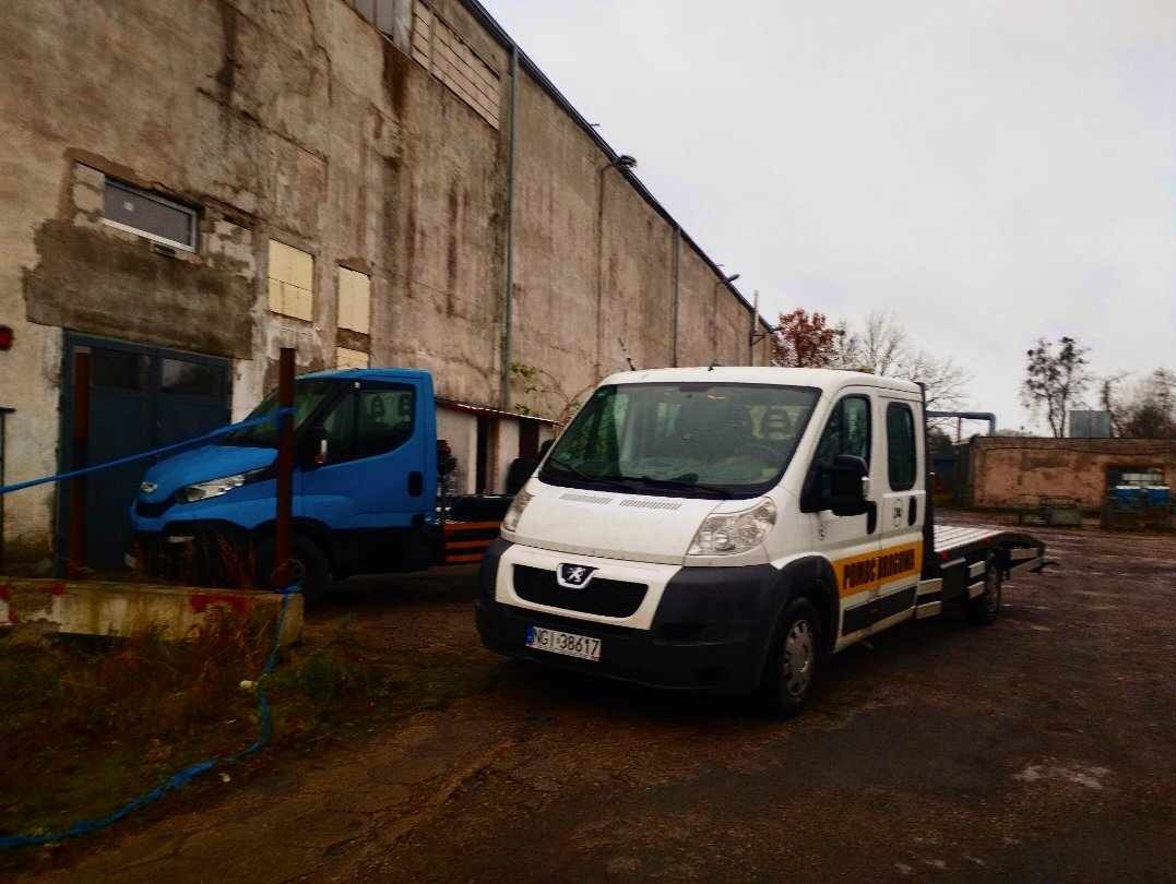 Dwa busy: biały bus laweta z żółtym napisem i niebieski bus, stojące przed szarym, betonowym budynkiem w pochmurny dzień.