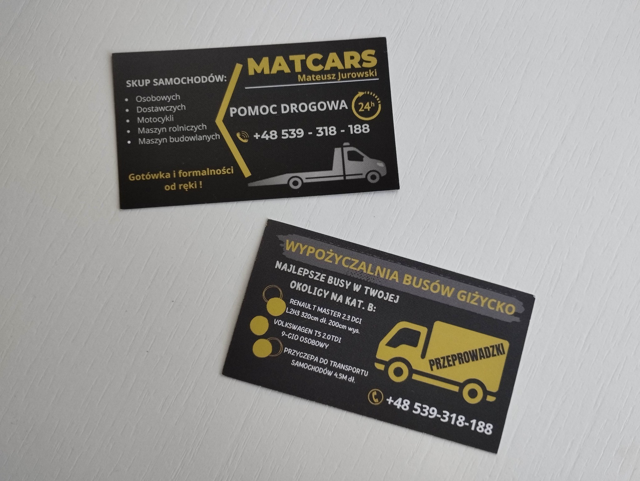 Dwie czarno-żółte wizytówki: jedna z ofertą skupu i pomocy drogowej, druga z wypożyczalnią busów w Giżycku, obie z numerem telefonu +48 539-318-188.