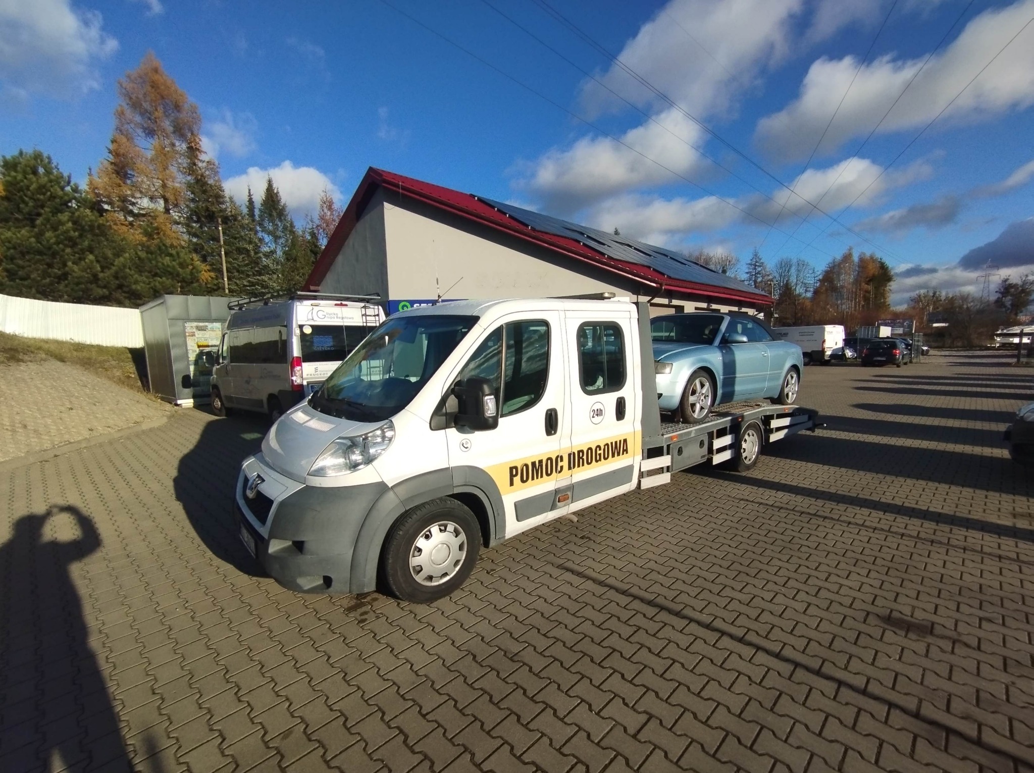 Laweta z błękitnym autem osobowym na platformie, zaparkowana na brukowanym placu przed budynkiem z panelami słonecznymi na dachu, w słoneczny dzień.