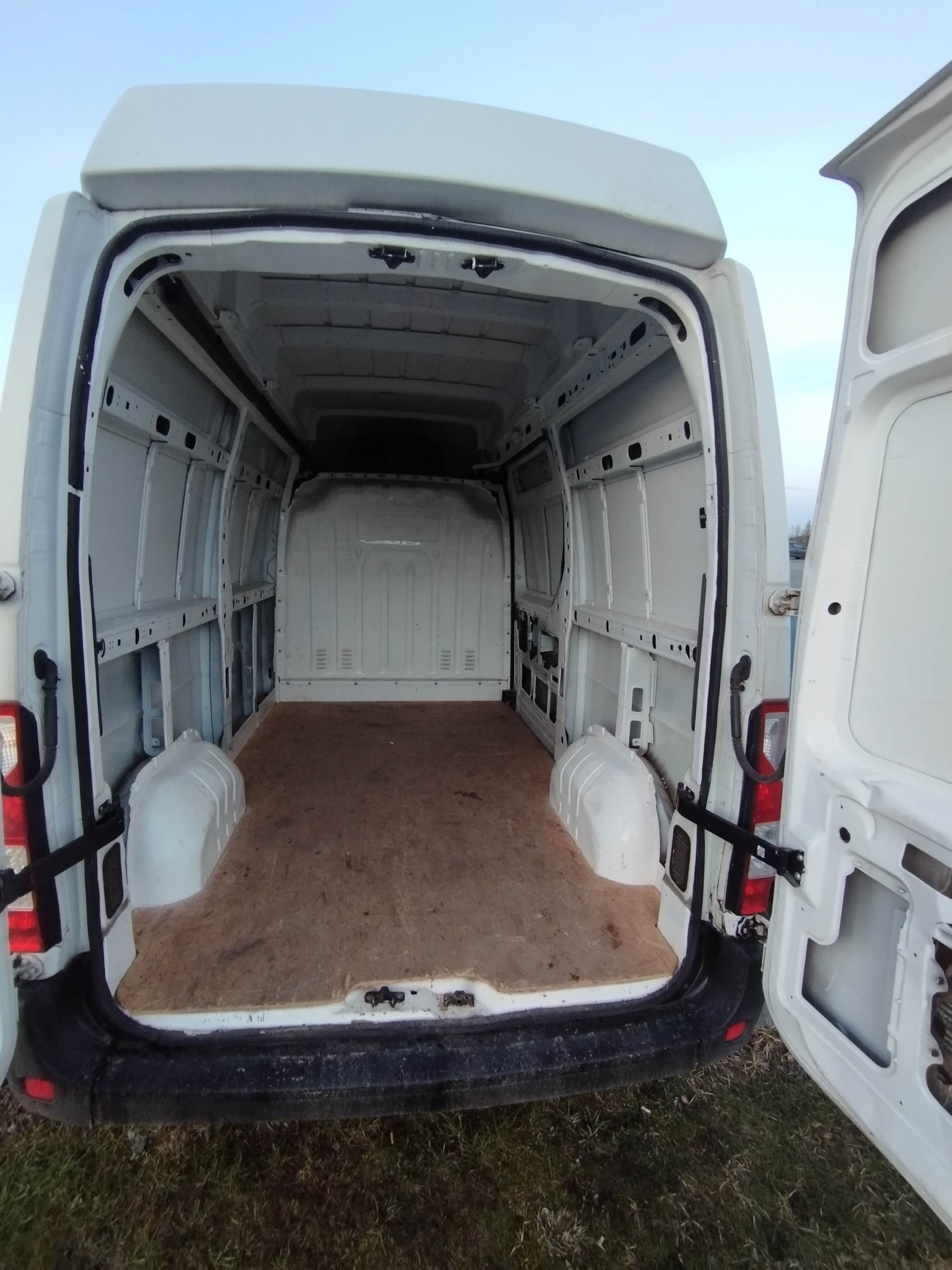 Wnętrze białego, pustego furgonu z otwartymi tylnymi drzwiami, ukazujące przestrzeń ładunkową z drewnianą podłogą i widocznymi elementami konstrukcyjnymi.