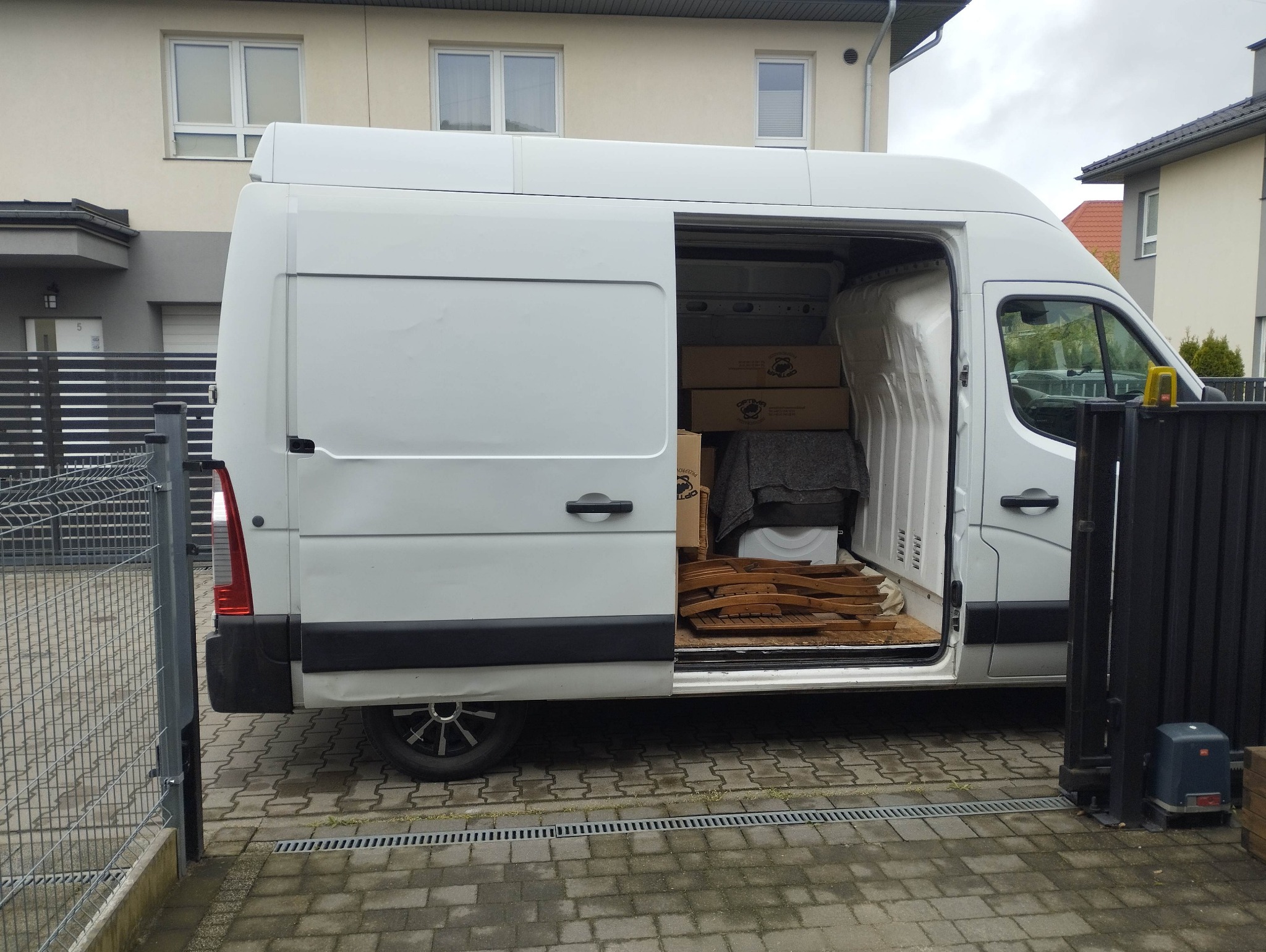 Biały van z otwartymi drzwiami bocznymi, częściowo załadowany kartonami i elementami drewnianymi, zaparkowany na posesji z metalowym ogrodzeniem.