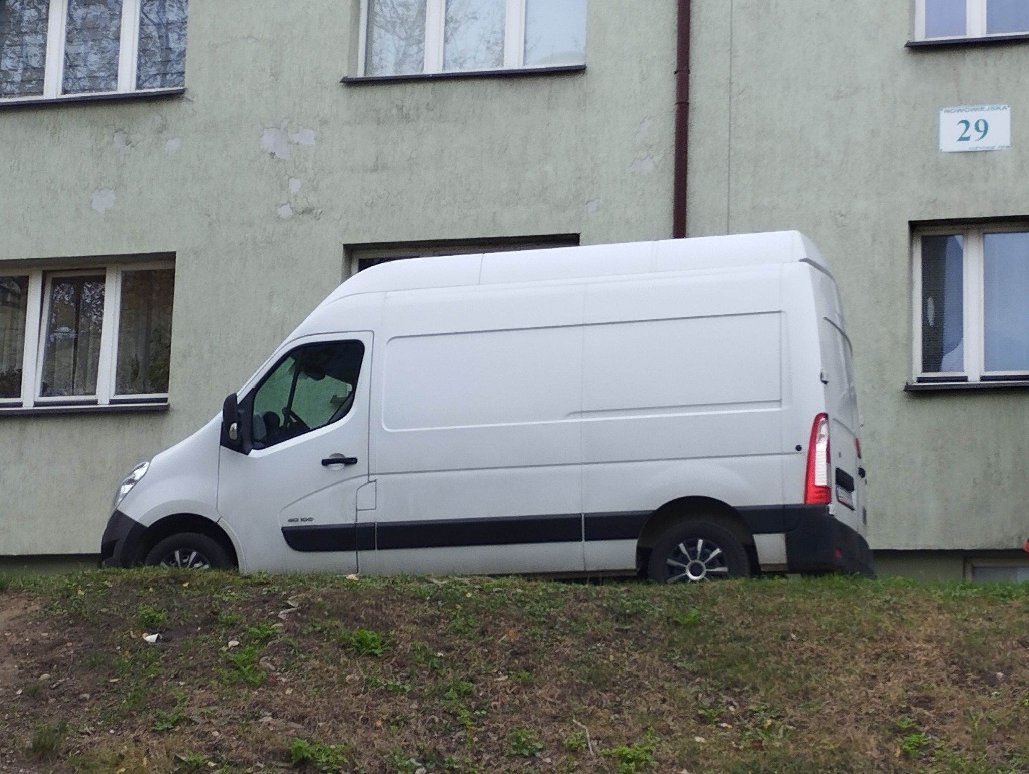 Biały furgon Renault Master zaparkowany na trawiastym wzgórzu przed budynkiem mieszkalnym z numerem 29, widok z boku, pochmurny dzień.