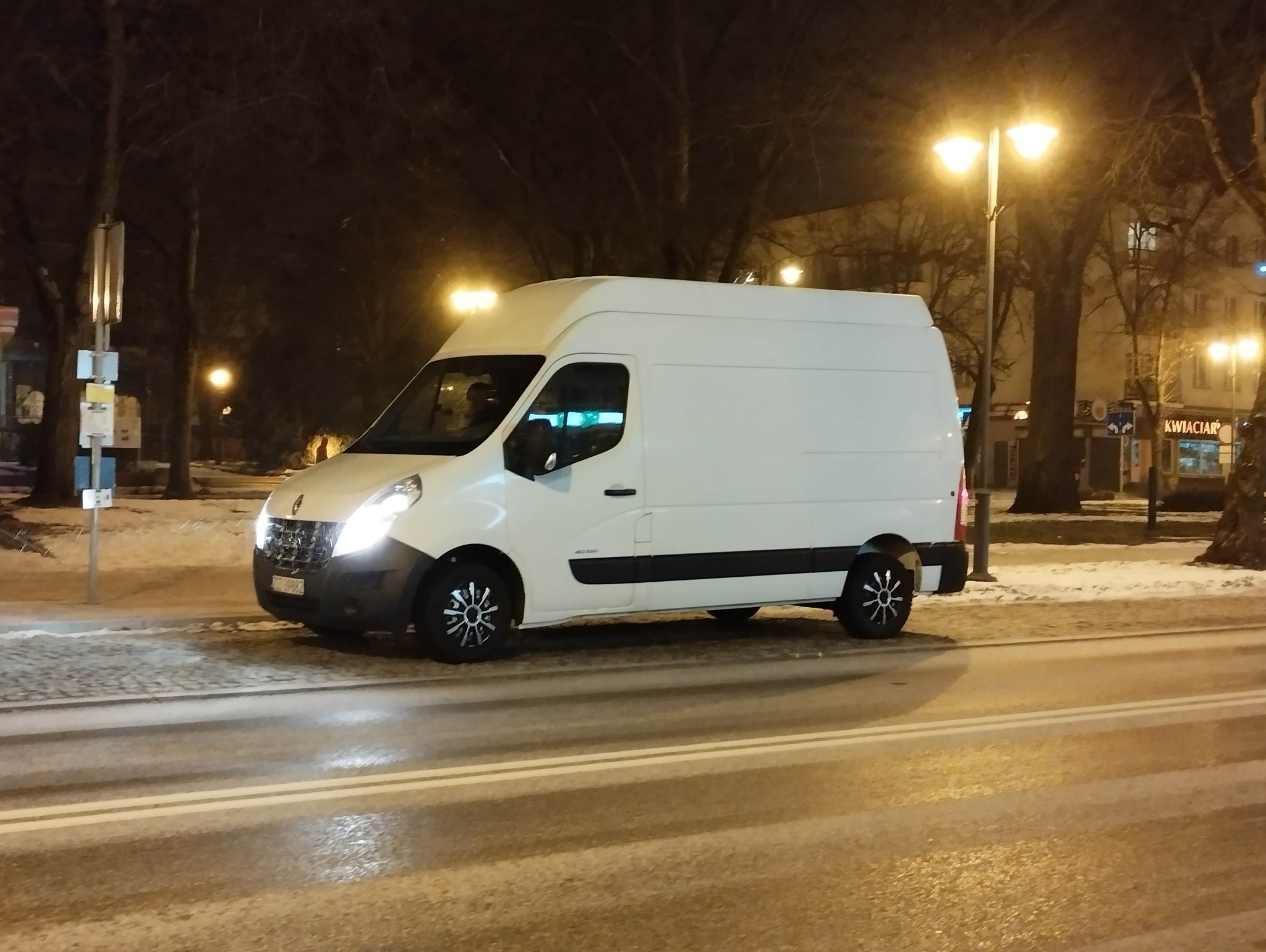 Biały bus Renault Master na tle nocnego, zimowego krajobrazu miejskiego. Widoczne oświetlenie uliczne i zasypane śniegiem pobocze.