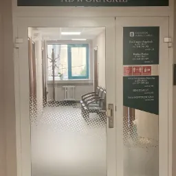 Wejście do kancelarii adwokackiej z widocznymi nazwiskami adwokatów na drzwiach i wnętrzem z wieszakiem na ubrania oraz krzesłami w tle.