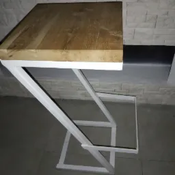 Blat z jasnego drewna na białym metalowym stelażu o geometrycznej konstrukcji, na tle ściany z imitacji kamienia.