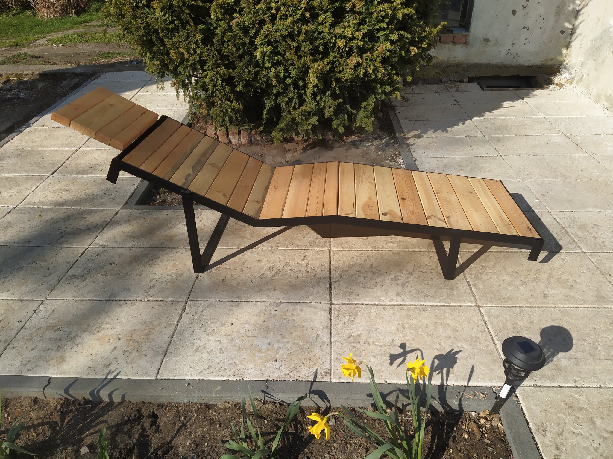 Drewniany leżak ogrodowy z czarną metalową ramą, ustawiony na płytach chodnikowych, obok rabaty z żółtymi narcyzami i lampą solarną.