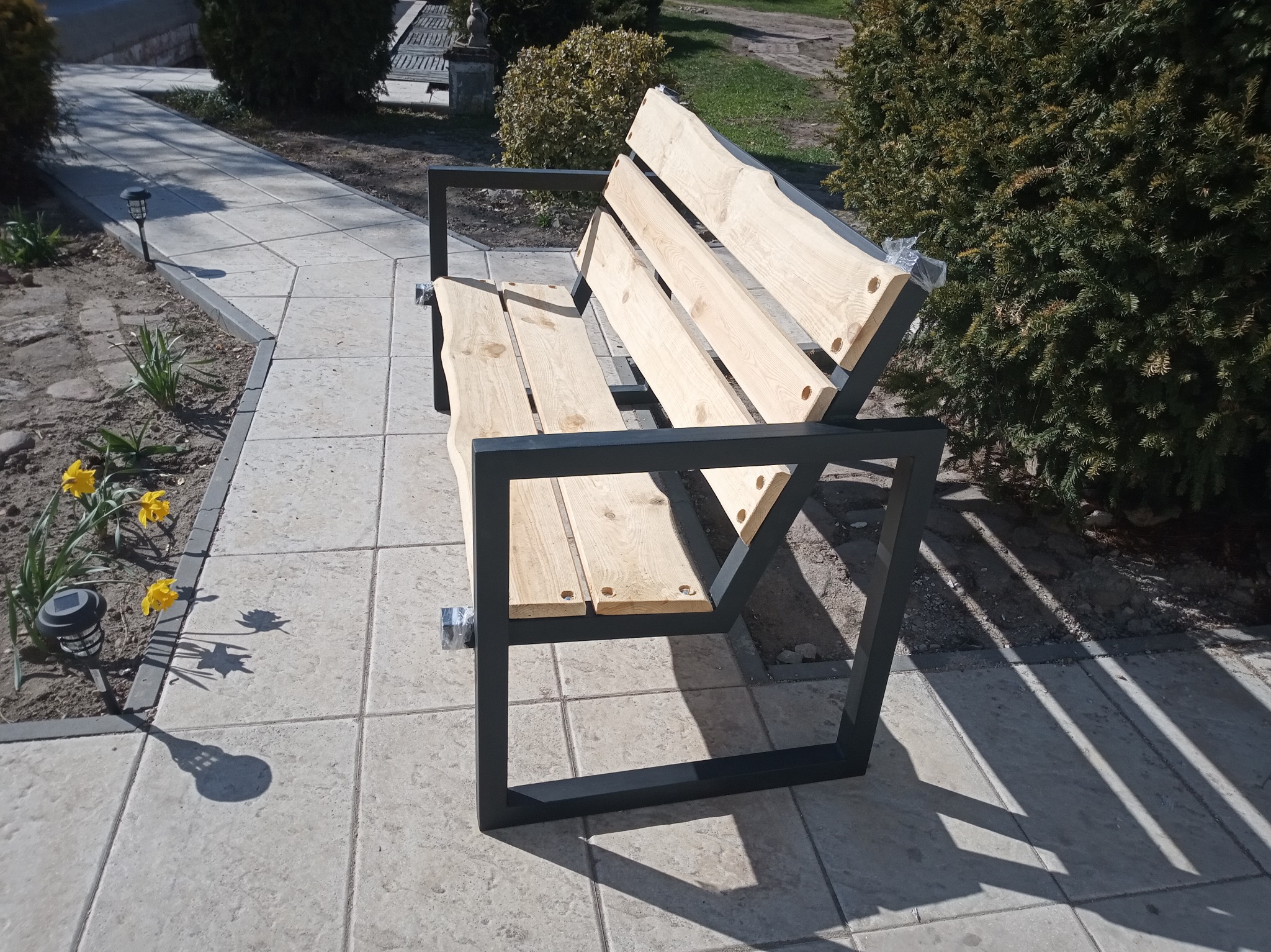 Nowoczesna ławka ogrodowa z surowego drewna i czarnej metalowej ramy, umieszczona na wyłożonym płytami chodniku, z widocznymi żółtymi kwiatami i krzewami w tle.