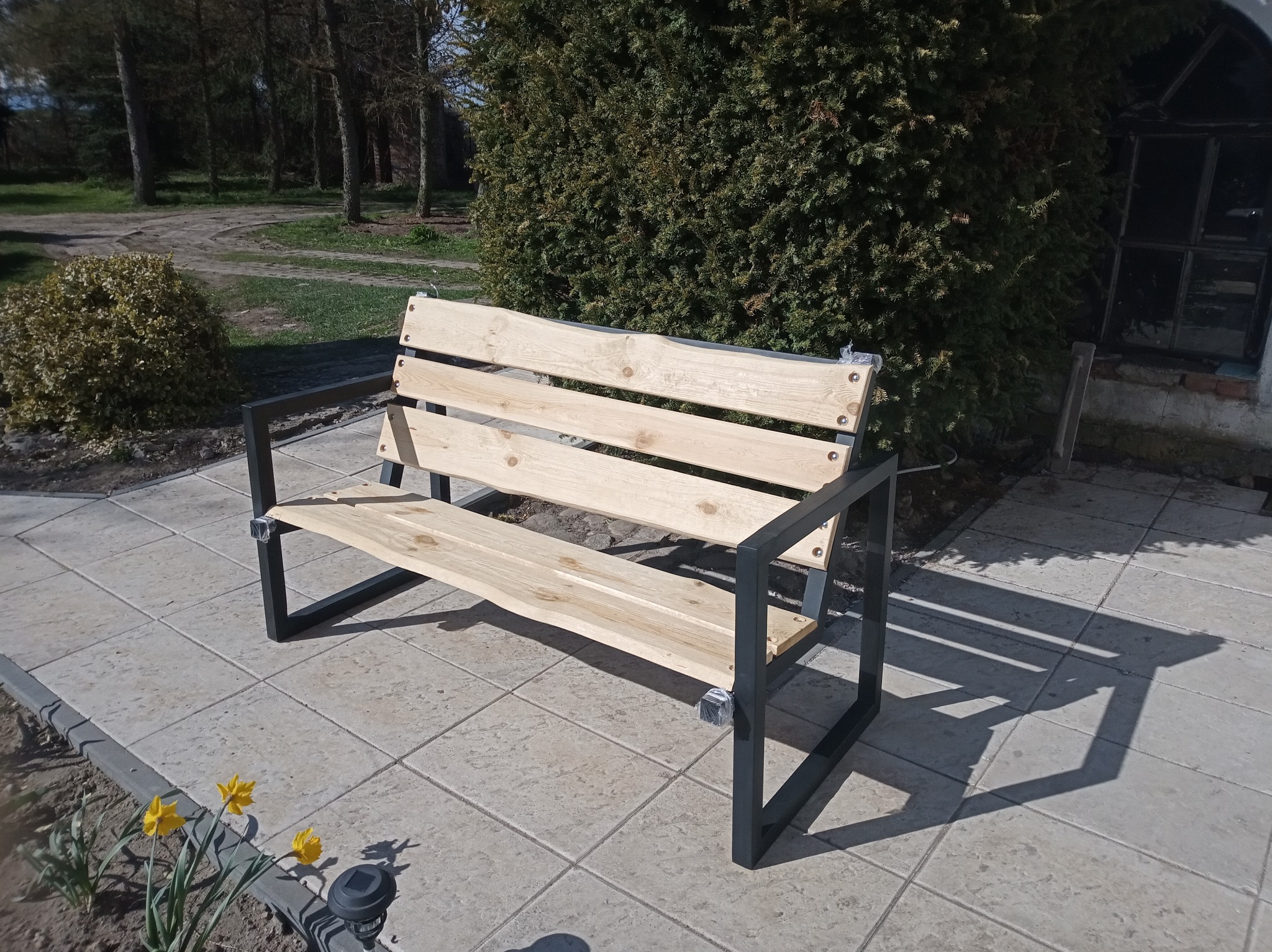 Nowoczesna ławka ogrodowa z jasnego drewna i czarnych metalowych nóg, ustawiona na płytkach chodnikowych w słoneczny dzień, z widocznymi żółtymi kwiatami w tle.