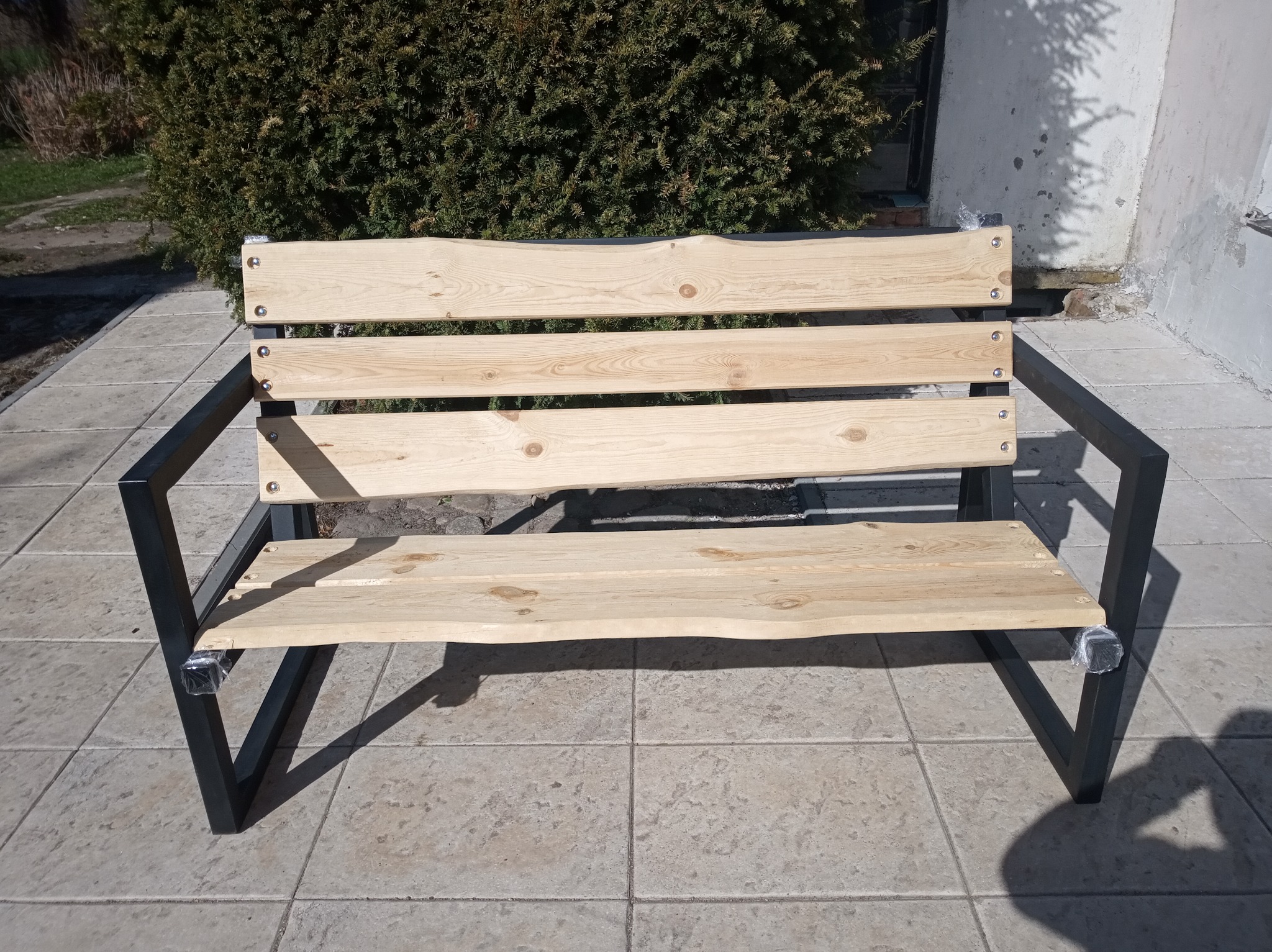 Surowa ławka ogrodowa z jasnego drewna i czarnych metalowych nóg, z zabezpieczeniami na stopkach, na tle kostki brukowej i zielonego krzewu.