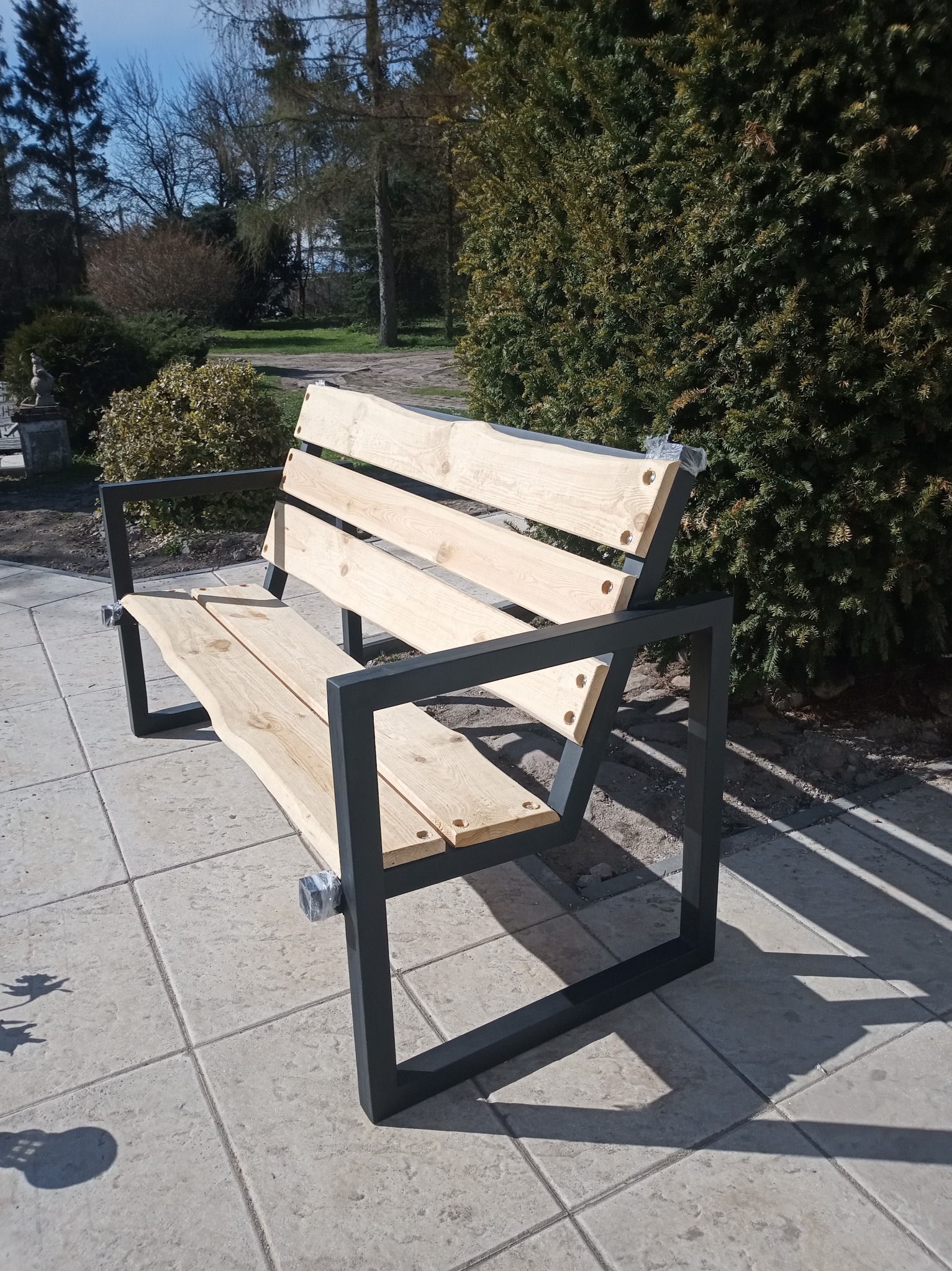 Nowoczesna ławka parkowa z surowego drewna i stalowej ramy, zabezpieczona folią ochronną na zakończeniach, na tle zieleni parkowej i chodnika z płyt.