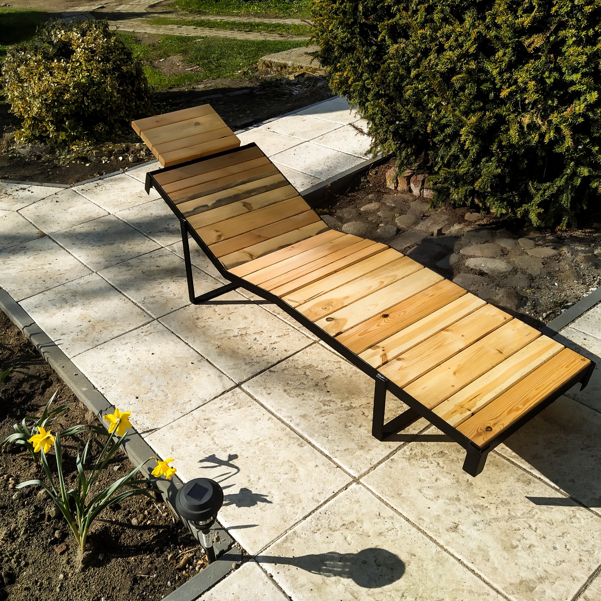 Drewniany leżak ogrodowy z czarną metalową ramą, ustawiony na jasnych płytach chodnikowych, obok rabaty z kwitnącymi na żółto narcyzami.