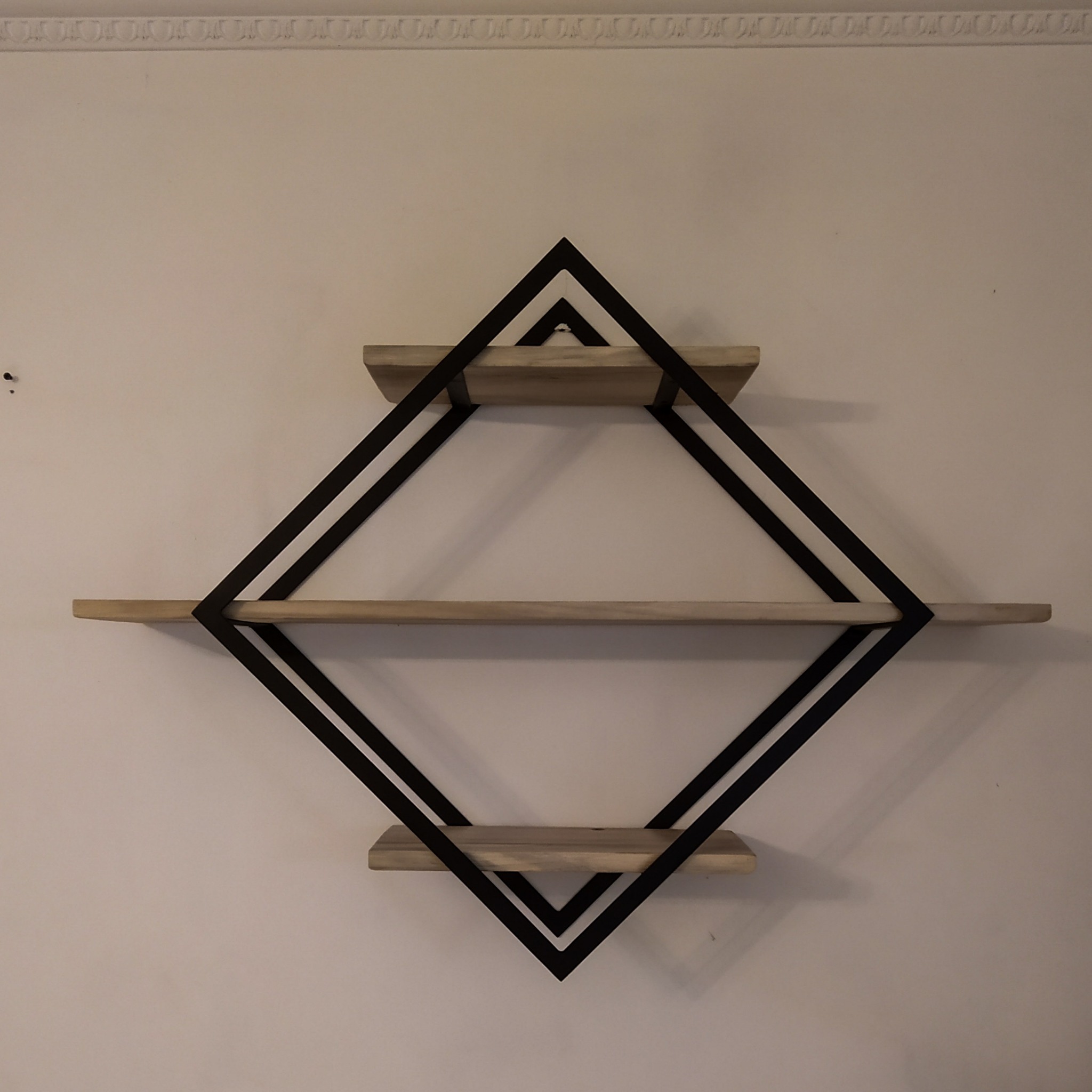 Półka ścienna z trzema drewnianymi blatami w geometrycznej, podwójnej ramie w kształcie rombu z czarnych płaskowników, zamocowana na jasnej ścianie z ozdobnym gzymsem.