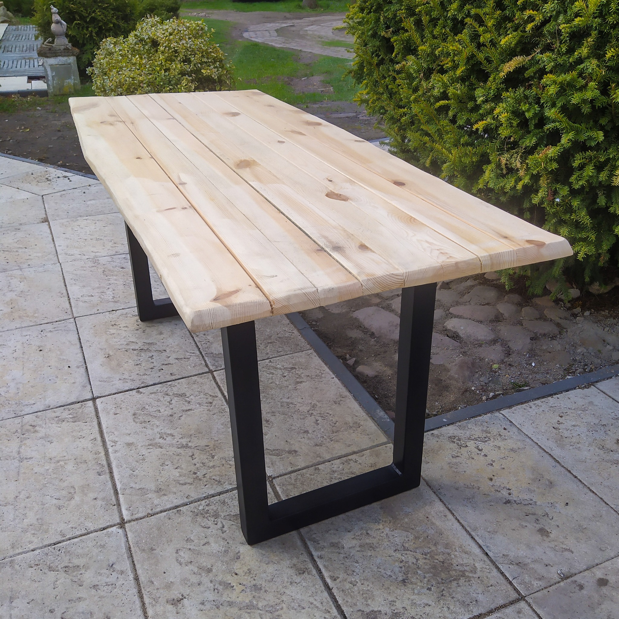 Surowy, jasny stół z desek z czarnymi, metalowymi nogami w kształcie litery U, ustawiony na kamiennej posadzce w ogrodzie.