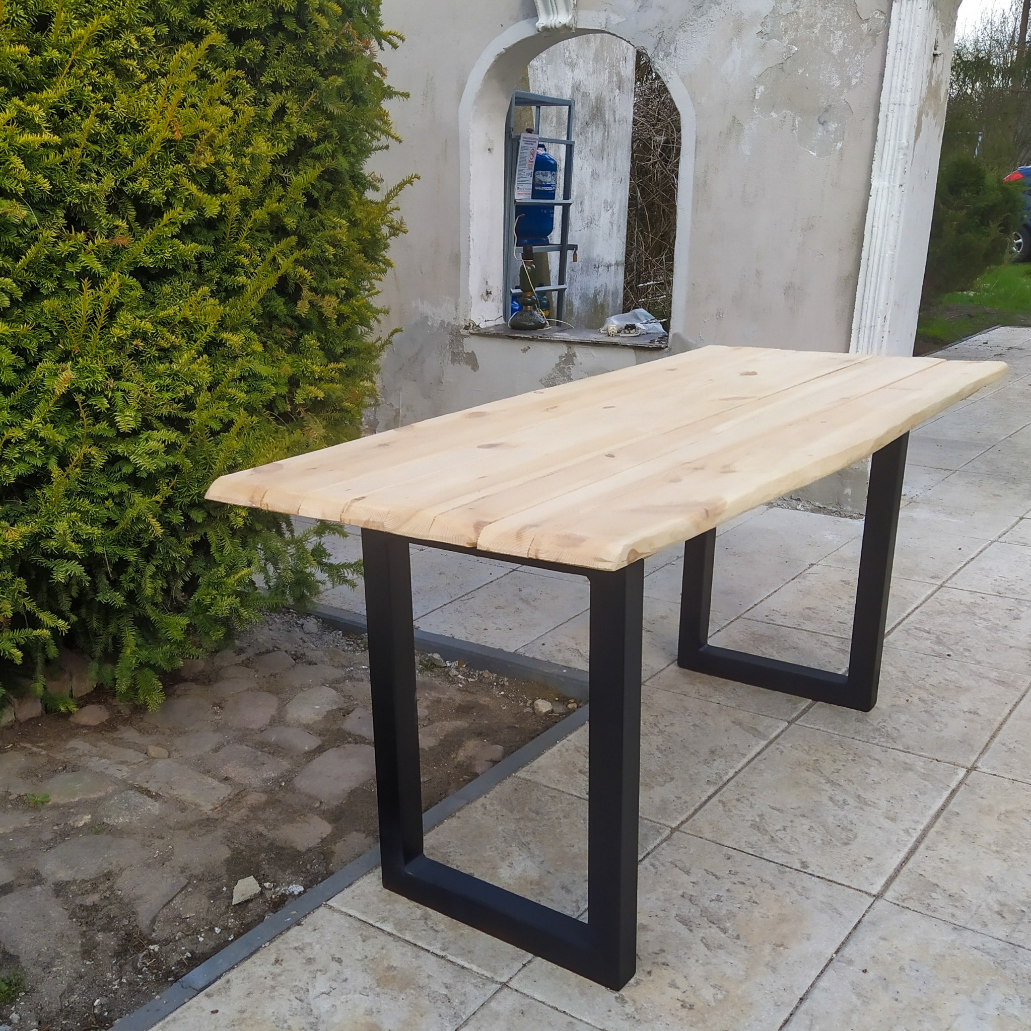 Surowy drewniany blat stołu na czarnych, metalowych nogach typu U, ustawiony na zewnątrz na tle krzewu i budynku w trakcie remontu.