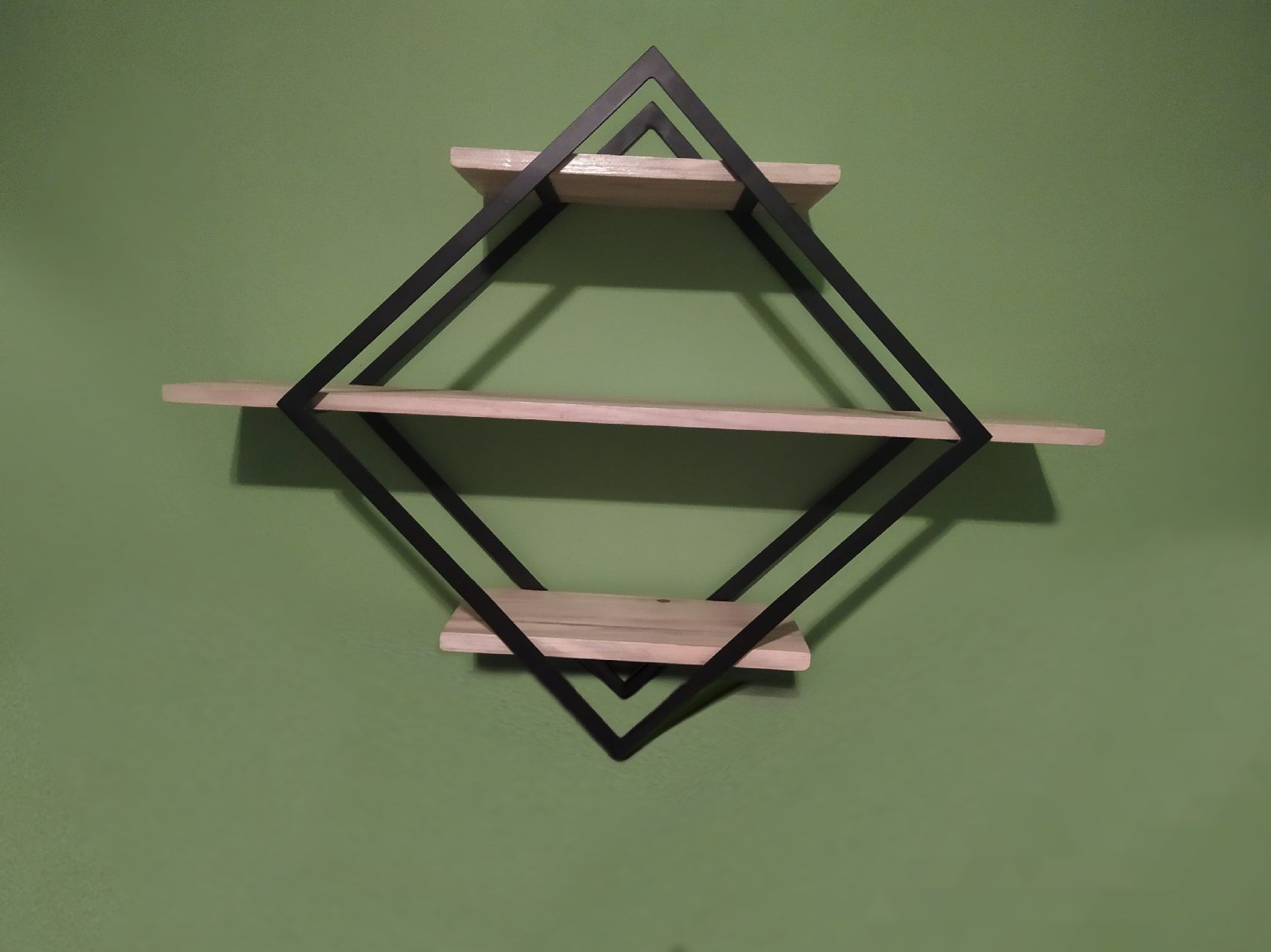 Półka ścienna z drewnianymi blatami i czarną, metalową ramą w kształcie rombu, na zielonej ścianie.