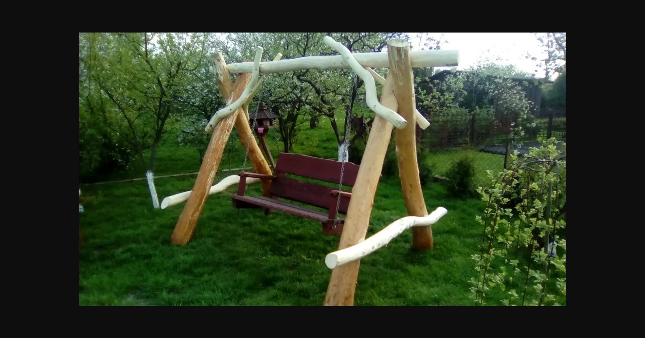 Drewniana huśtawka ogrodowa z naturalnych pni drzew, zawieszona na łańcuchach, z małym karmnikiem dla ptaków w tle, otoczona zielonym trawnikiem i kwitnącymi drzewami owocowymi.