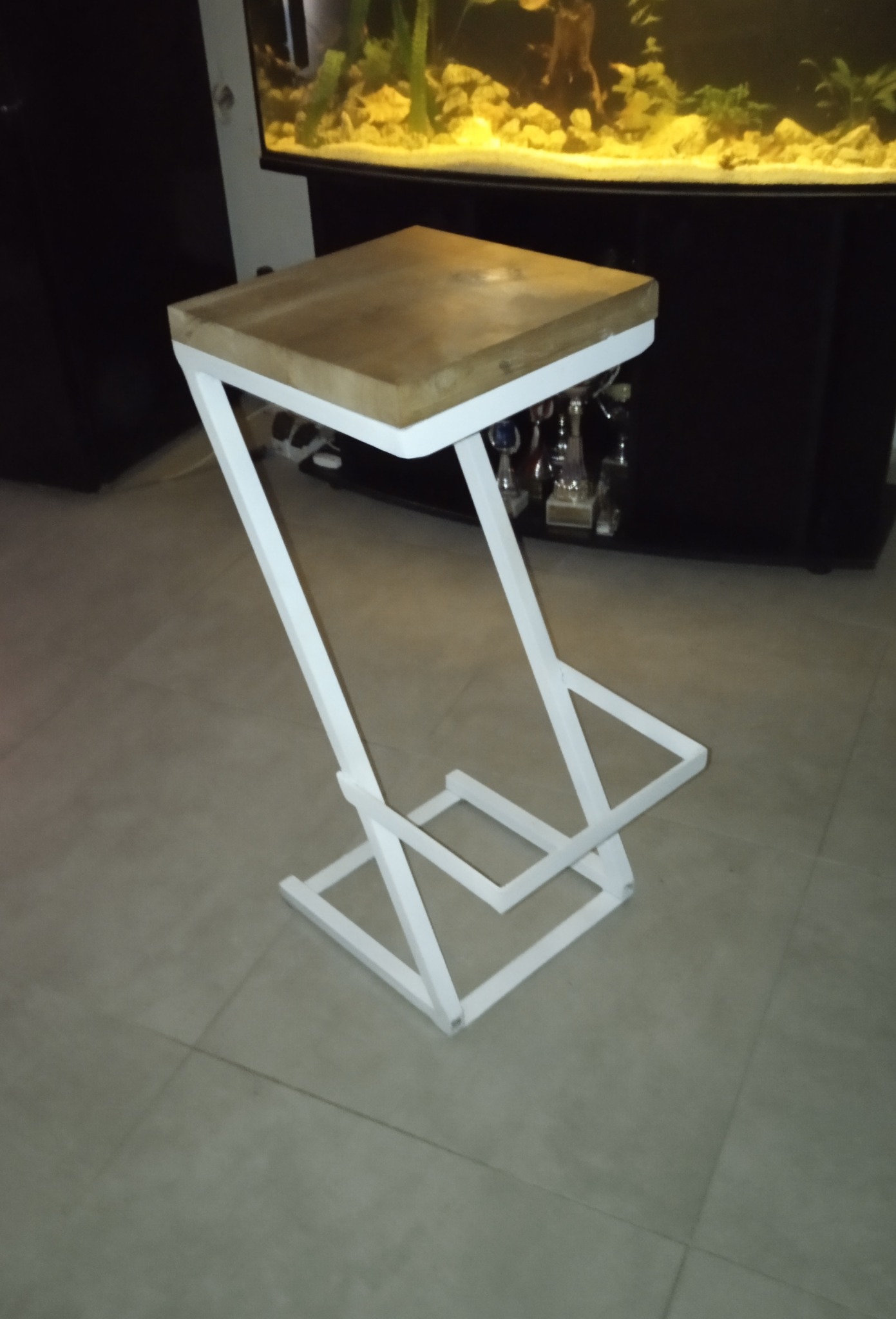 Nowoczesny, minimalistyczny taboret z drewnianym blatem i białą, geometryczną podstawą z metalu, stoi na szarej podłodze, a w tle widoczny jest akwarium.