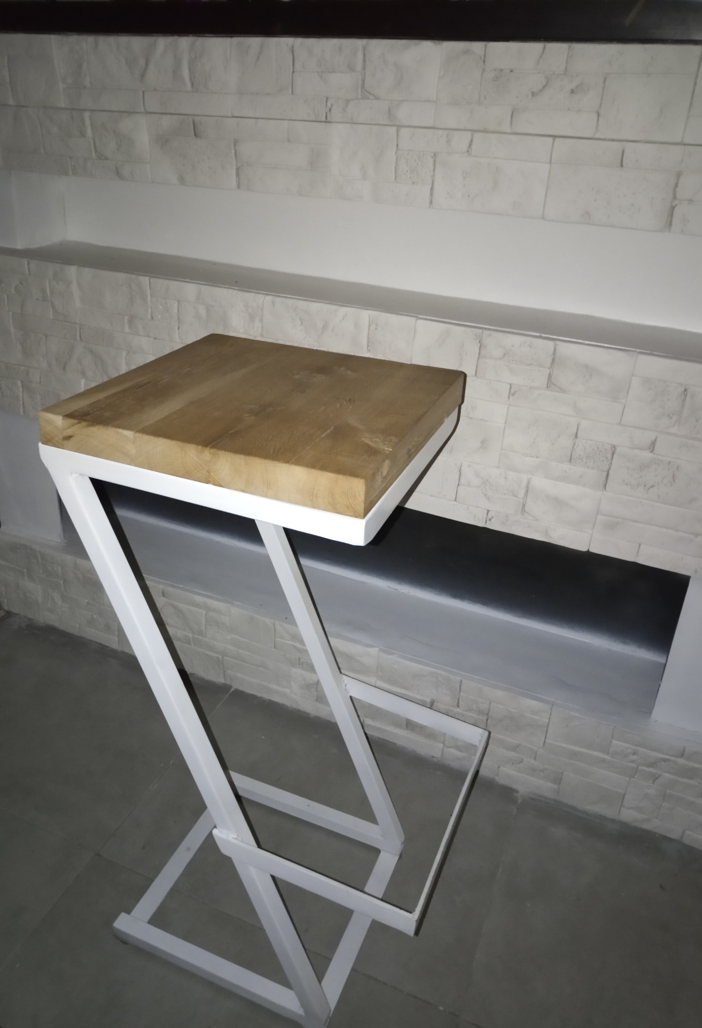 Nowoczesny stolik pomocniczy z blatem z jasnego drewna i białą, geometryczną podstawą z metalu na tle ściany wyłożonej dekoracyjną cegłą.