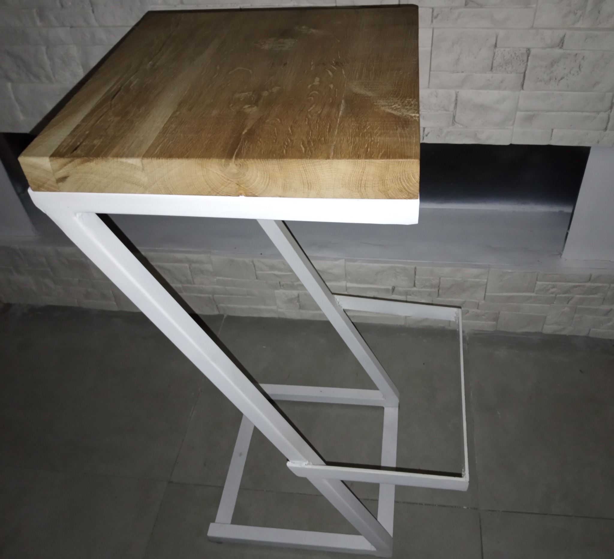 Blat z jasnego drewna na białym metalowym stelażu o geometrycznej konstrukcji, na tle ściany z imitacji kamienia.