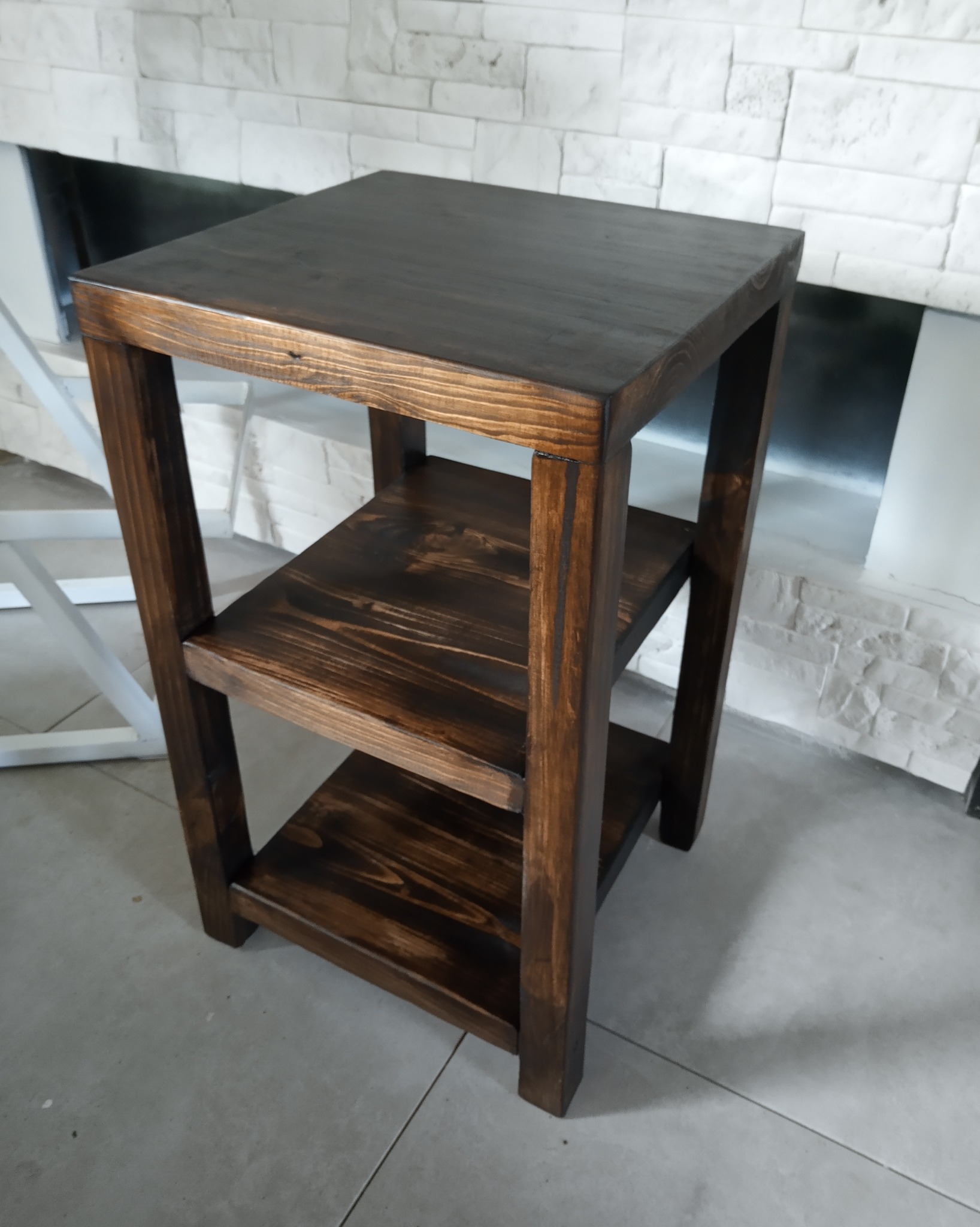 Drewniany stolik nocny z trzema półkami, bejcowany na ciemny brąz, na tle ściany z białej cegły.