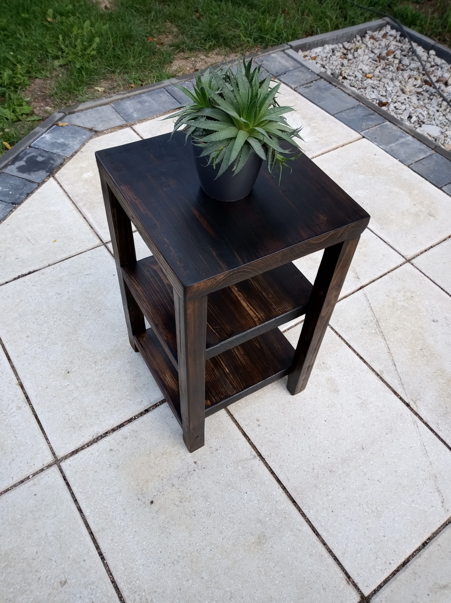 Ciemny, drewniany stolik z półkami na zewnątrz, na którym stoi doniczka z zieloną rośliną, na tle jasnej kostki brukowej i trawy.