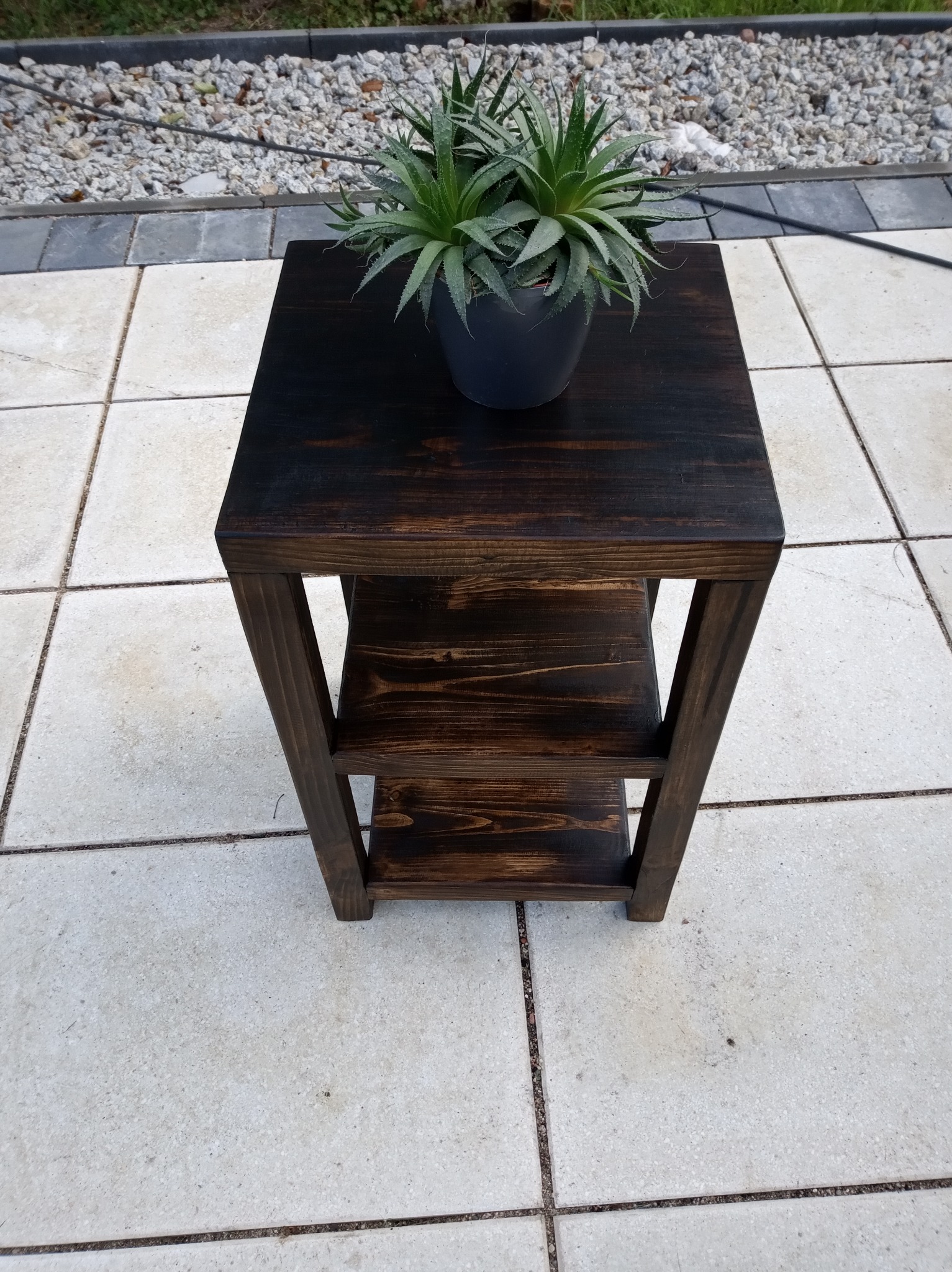 Drewniany stolik z dwiema półkami, bejcowany na ciemny brąz, z rośliną doniczkową na blacie, ustawiony na jasnych płytach tarasowych.