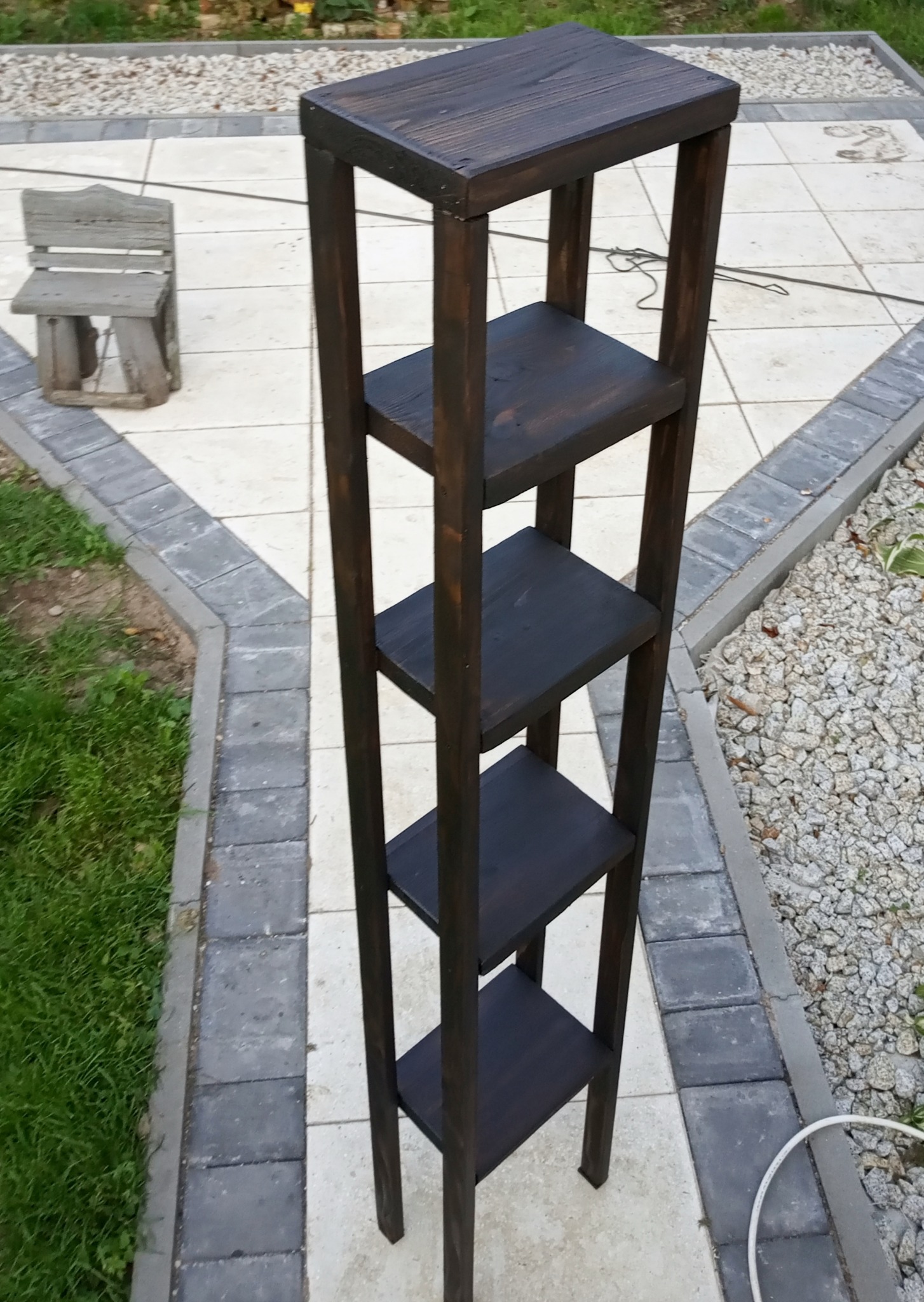 Wysoki, wąski regał z ciemnego drewna, z czterema półkami, stojący na zewnątrz na płytkach chodnikowych, obok trawnika i kamieni ozdobnych.