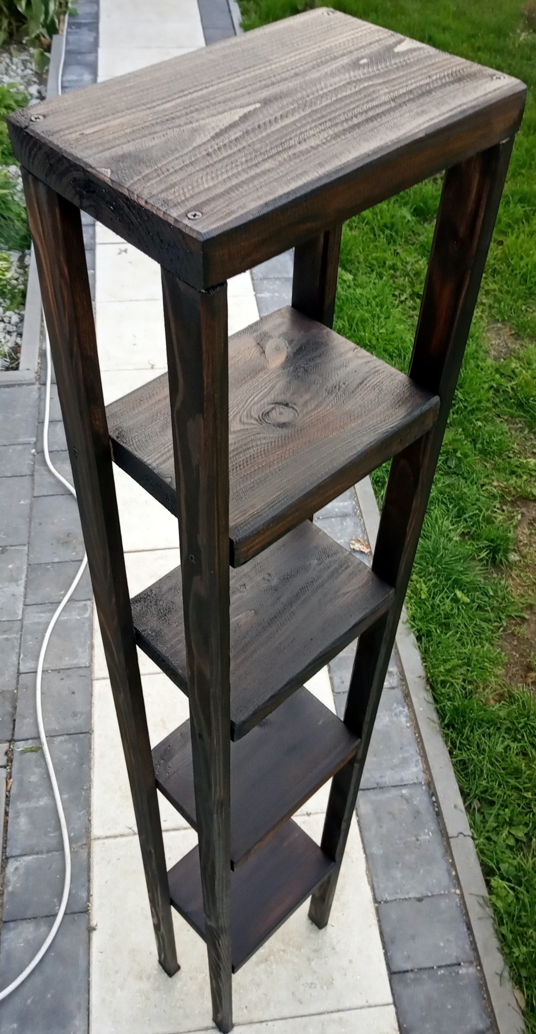 Ciemnobrązowy, drewniany regał drabinkowy z czterema półkami na tle kostki brukowej i trawy.