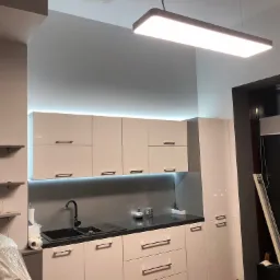 Nowoczesna kuchnia z białymi szafkami, czarnym blatem i minimalistycznym oświetleniem LED pod szafkami wiszącymi, widoczna instalacja elektryczna w postaci gniazdka na blacie.