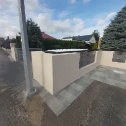 Nowoczesny mur z elementami metalowymi, wykończony białymi kamieniami, otoczony szarą kostką brukową, widok na domy w tle pod błękitnym niebem.