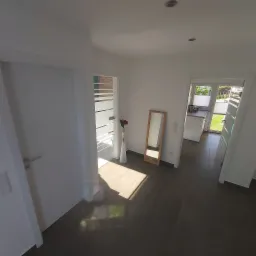 Jasne wnętrze domu po remoncie: ciemna podłoga z płytek, białe ściany i drzwi, wysokie lustro w drewnianej ramie oraz wazon z kwiatami.