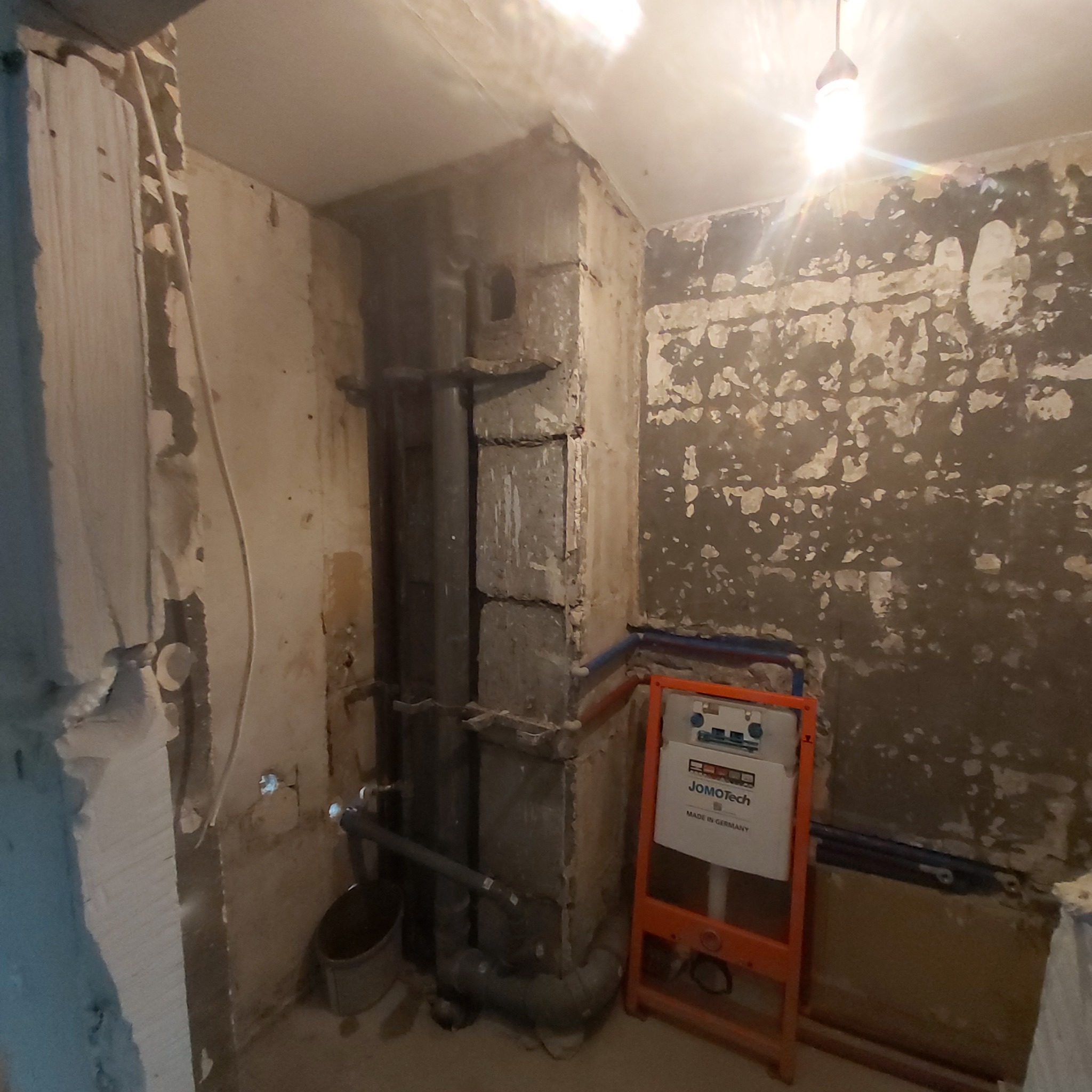 Demontaż starej instalacji w łazience: odsłonięte rury kanalizacyjne, fragment ściany z resztkami starej glazury, stelaż podtynkowy WC JomoTech przed montażem.