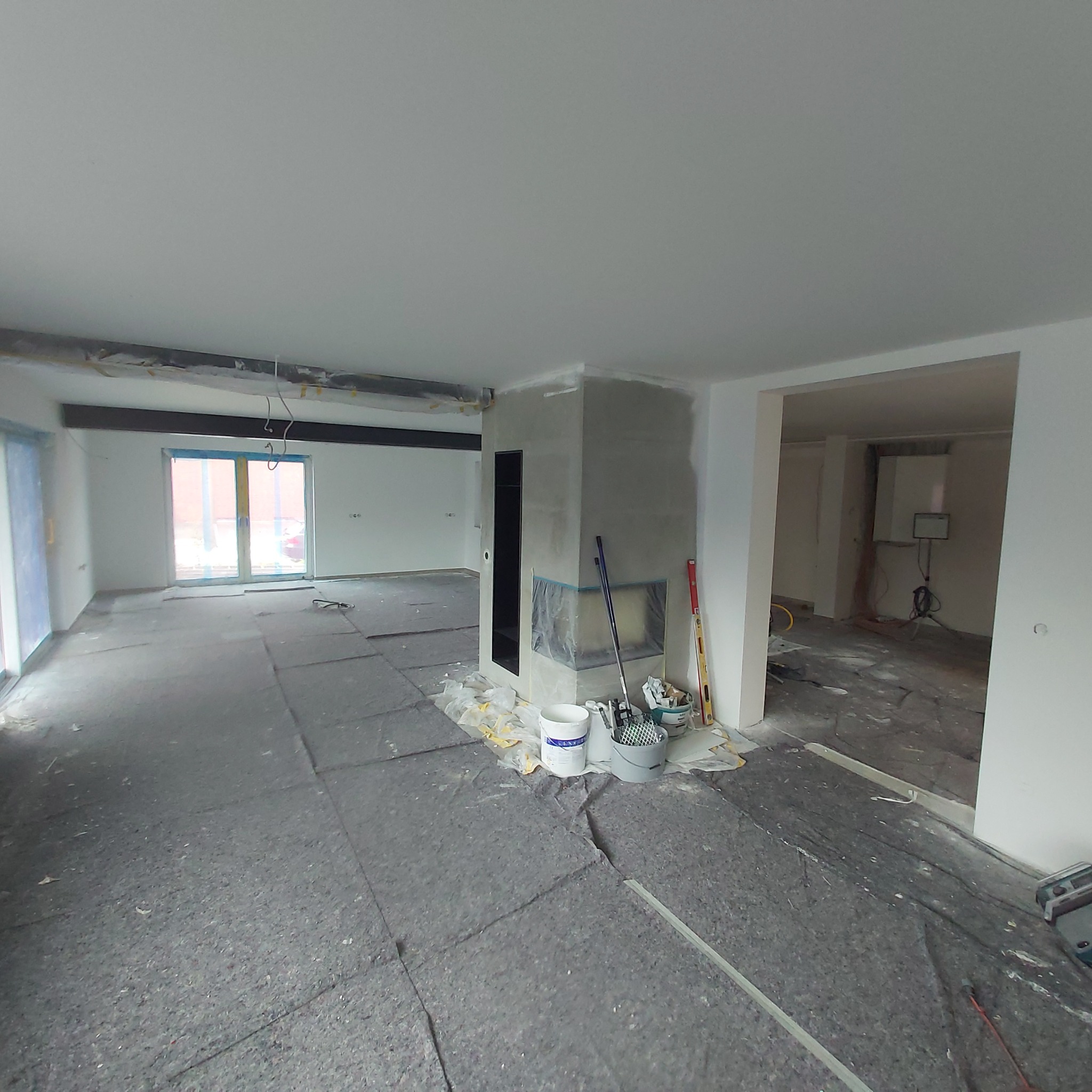 Wnętrze w trakcie remontu: zabezpieczona podłoga, ściany w trakcie gładzenia, zabudowa kominkowa przed wykończeniem, widoczne narzędzia i materiały budowlane.