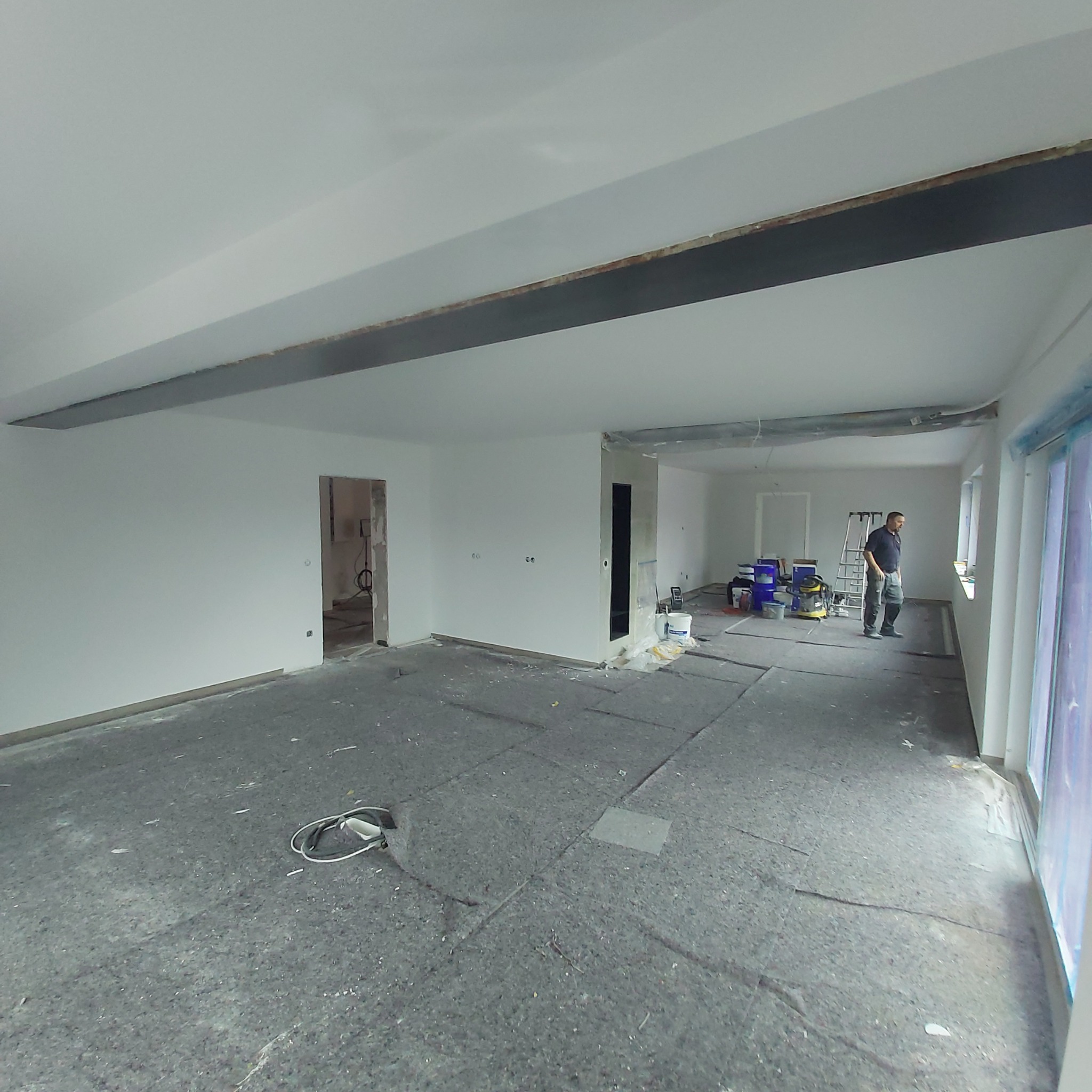 Przestronne wnętrze w trakcie malowania ścian na biało; widoczny pracownik, drabina, wiaderka z farbą oraz zabezpieczone okna folią.