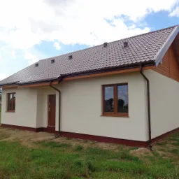 Niski, parterowy dom z brązowym dachem z blachodachówki, beżową elewacją i brązowymi oknami, wykończony drewnianą szczytową ścianą, w otoczeniu trawy i drzew