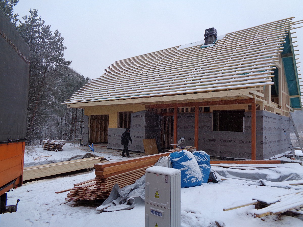 Budowa domu w zimowej scenerii: widoczne elementy konstrukcji szkieletowej, przygotowane drewno, folia izolacyjna Corotop i skrzynka elektryczna z ostrzeżeniami.