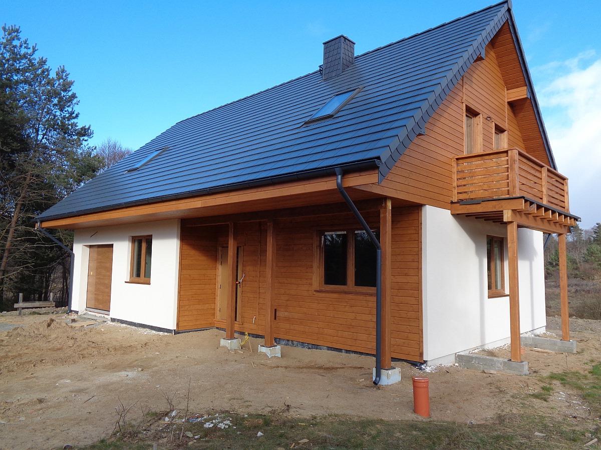 Nowoczesny dom z drewnianą elewacją i balkonem w Rumi, z ciemną dachówką i białymi fragmentami ścian, widziany z zewnątrz na etapie prac wykończeniowych.