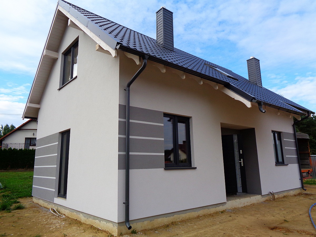 Nowoczesny dom z szarą elewacją z poziomymi paskami, ciemną dachówką i grafitowymi oknami, widok z boku na tle błękitnego nieba.