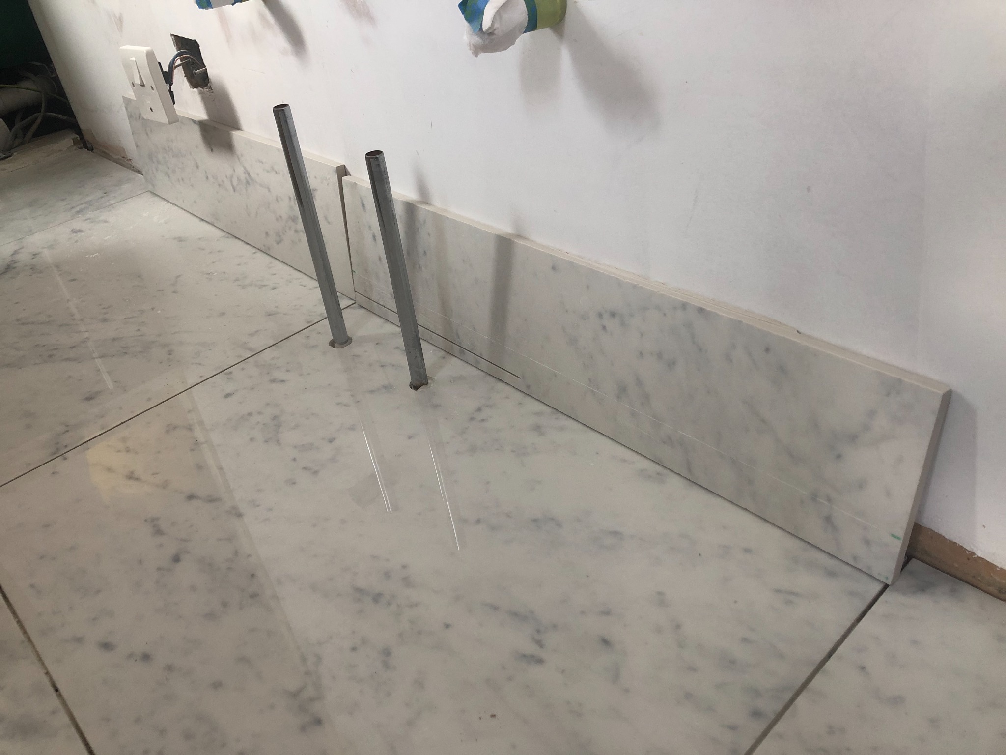 Montaż marmurowych płytek podłogowych i listew przypodłogowych w łazience, widoczne metalowe wsporniki i gniazdko elektryczne na ścianie.