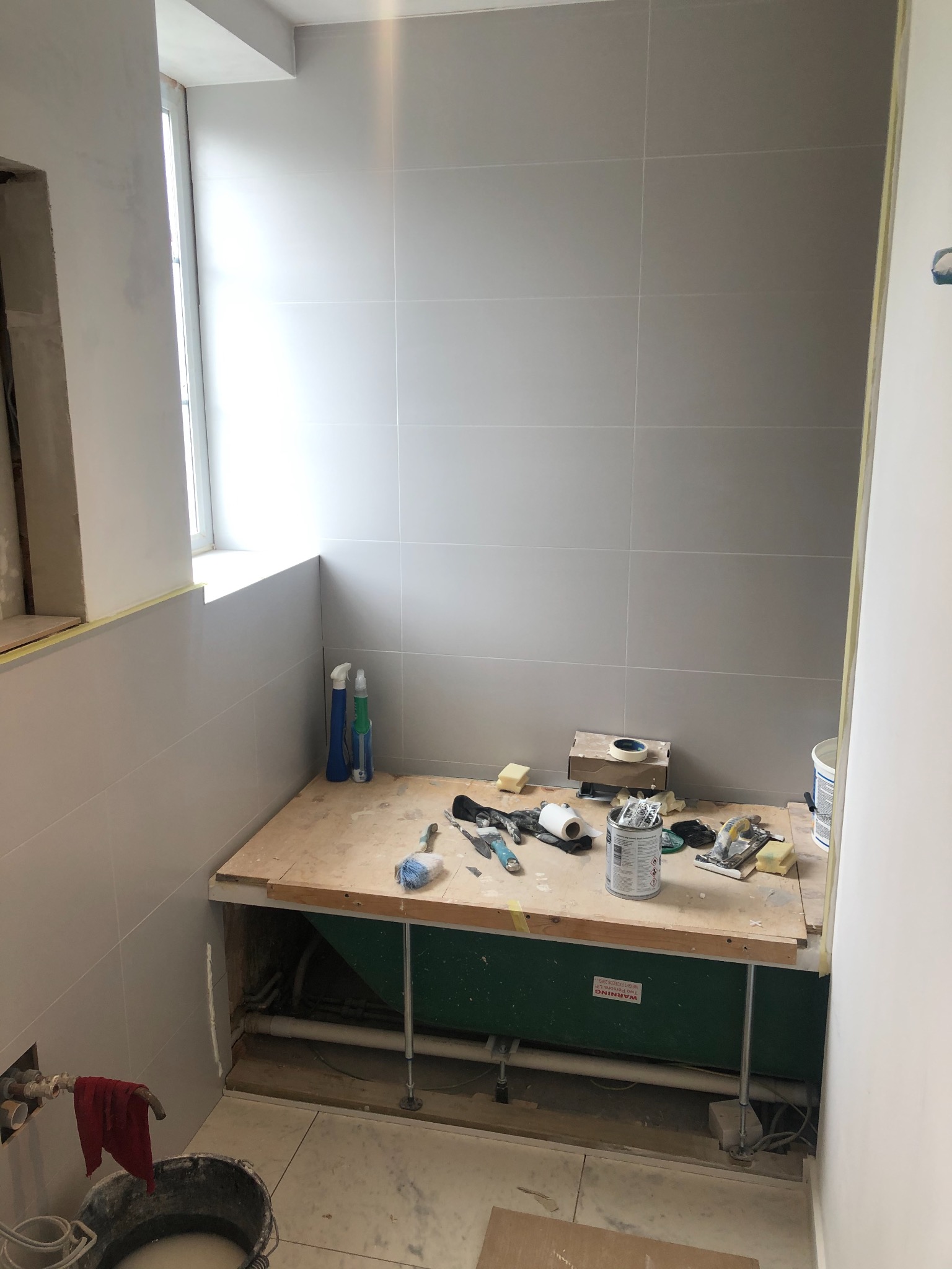 Remont łazienki w trakcie: szare płytki na ścianach, przygotowana wanna z obudową, narzędzia i materiały budowlane na drewnianej platformie, widoczne rury i instalacje, lekki nieporządek na podłodze.