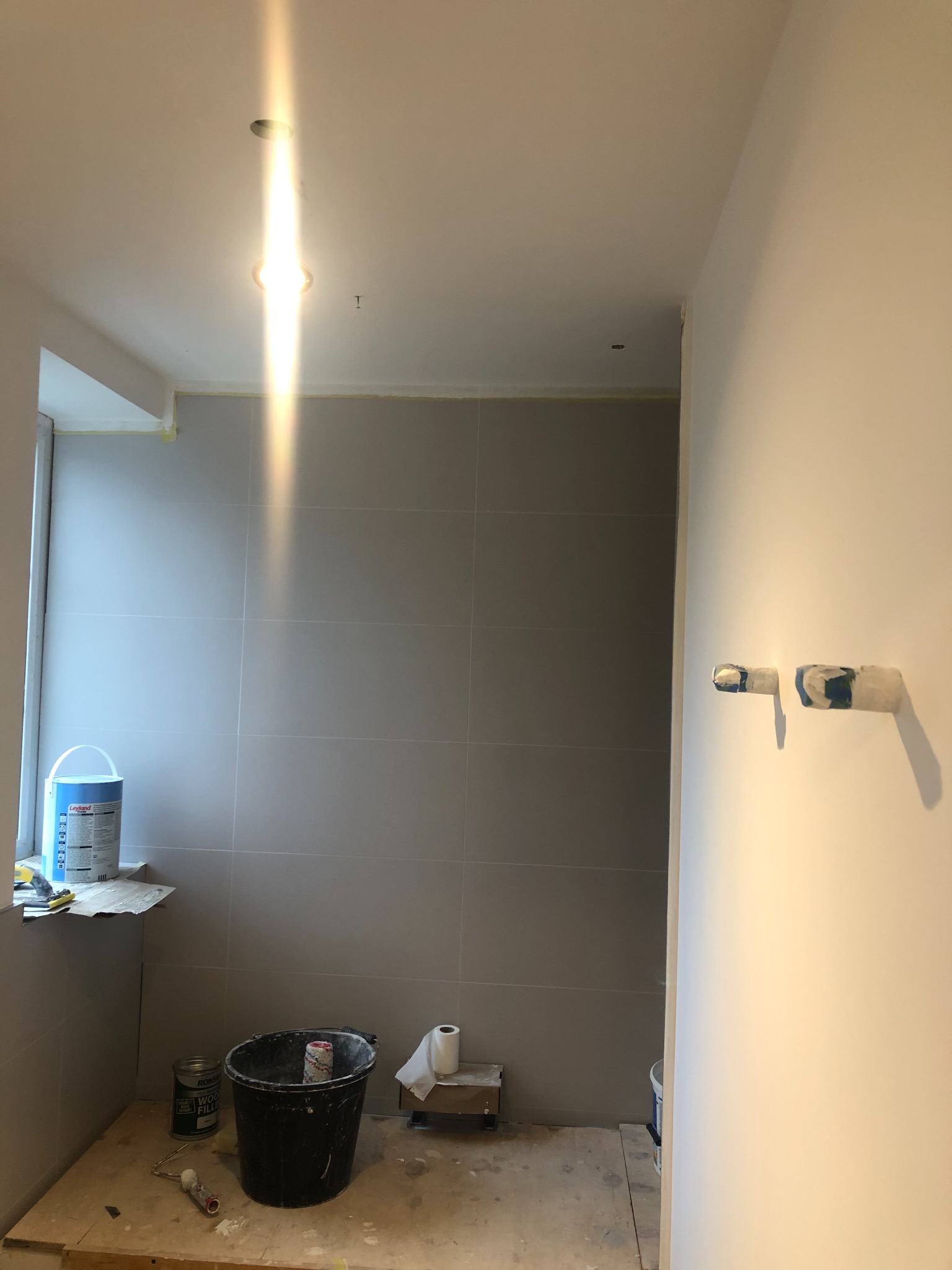 Wnętrze łazienki w trakcie prac wykończeniowych: szare płytki na ścianach, zabezpieczone taśmą malarską, widoczne narzędzia i materiały budowlane, przygotowane do malowania.