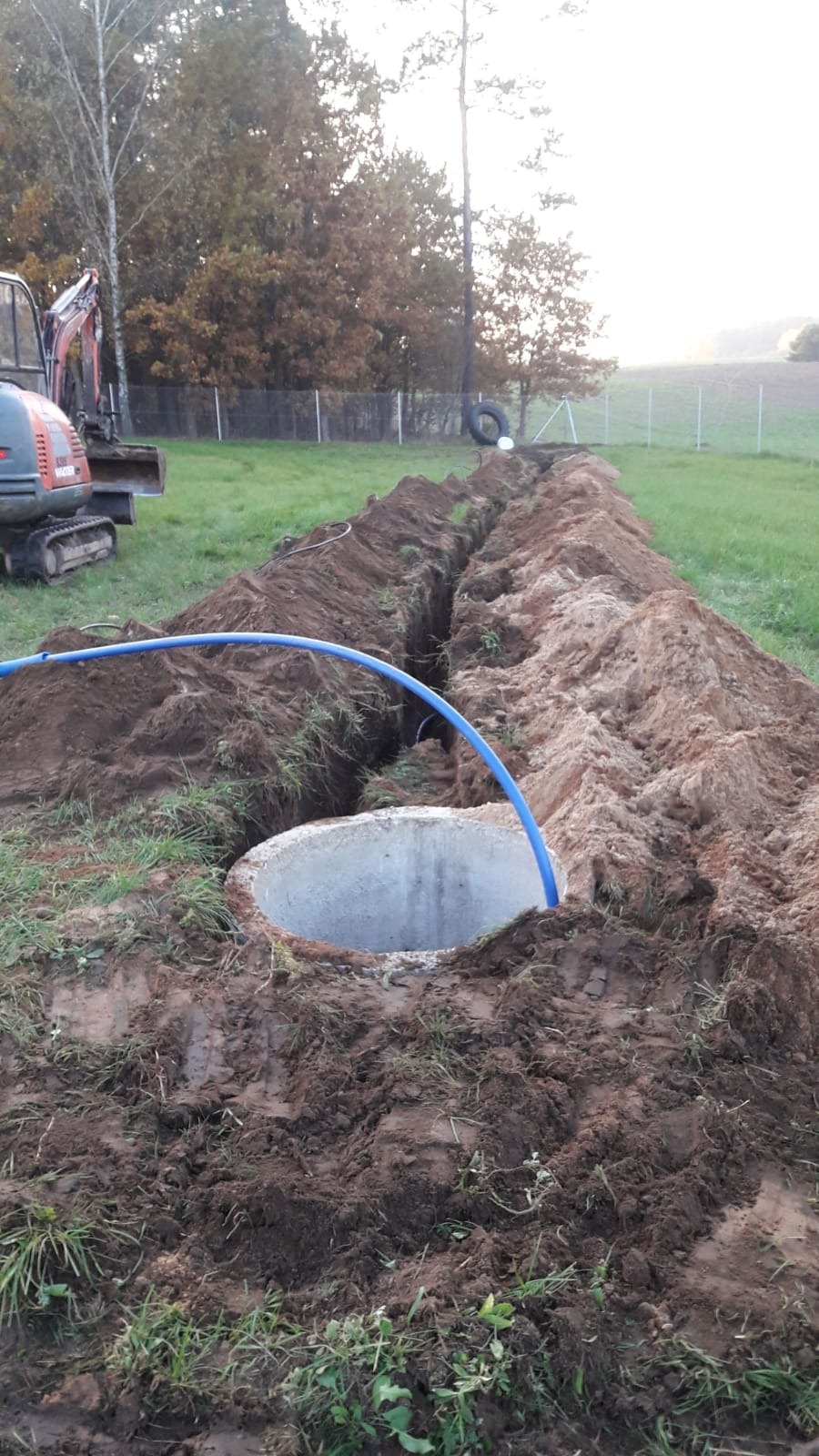 Wykonany wykop pod rurę doprowadzającą do betonowej studni, widoczna koparka w tle, teren trawiasty.