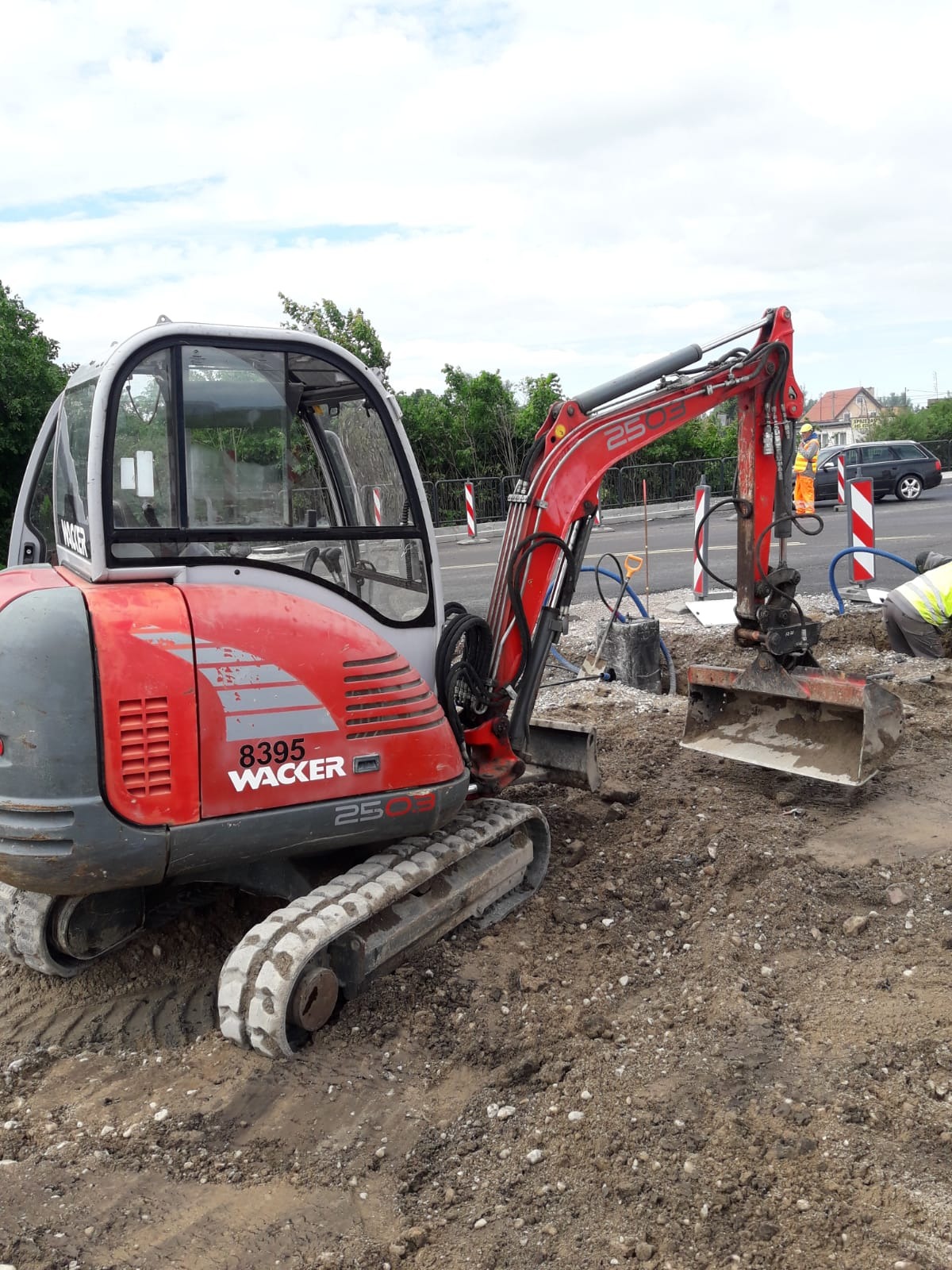 Czerwono-szara mini koparka Wacker 2503 na gąsienicach, z łyżką zanurzoną w ziemi, pracująca przy drodze; w tle widoczni robotnicy w odblaskowych ubraniach i zapory drogowe.