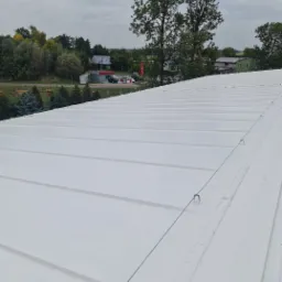 Świeżo pomalowany na biało dach z blachy trapezowej, widoczne linki zabezpieczające, w tle zabudowania wiejskie pod pochmurnym niebem.