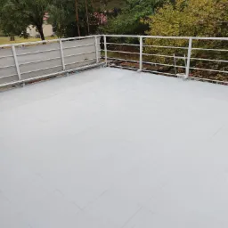 Biała, kwadratowa podłoga balkonu wykończona płytkami, otoczona szarą osłoną przeciwwiatrową i białą metalową barierką, z widokiem na drzewa w tle.