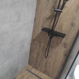 karmarRemonty Marcin Pański - Kabina prysznicowa walk-in z odpływem liniowym, wykończona płytkami imitującymi drewno na ścianie i podłodze, zestaw prysznicowy w kolorze czarnym, ściana wyłożona szarymi płytkami.
