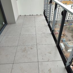 karmarRemonty Marcin Pański - Balkon z jasnymi płytkami podłogowymi, śladami brudu i częściowo odklejoną folią ochronną na szklanej balustradzie, z widokiem na zabudowę miejską w tle.